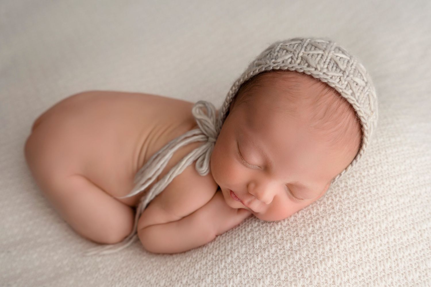 A sleeping newborn baby wearing a knit hat lies on soft white fabric in a peaceful pose.