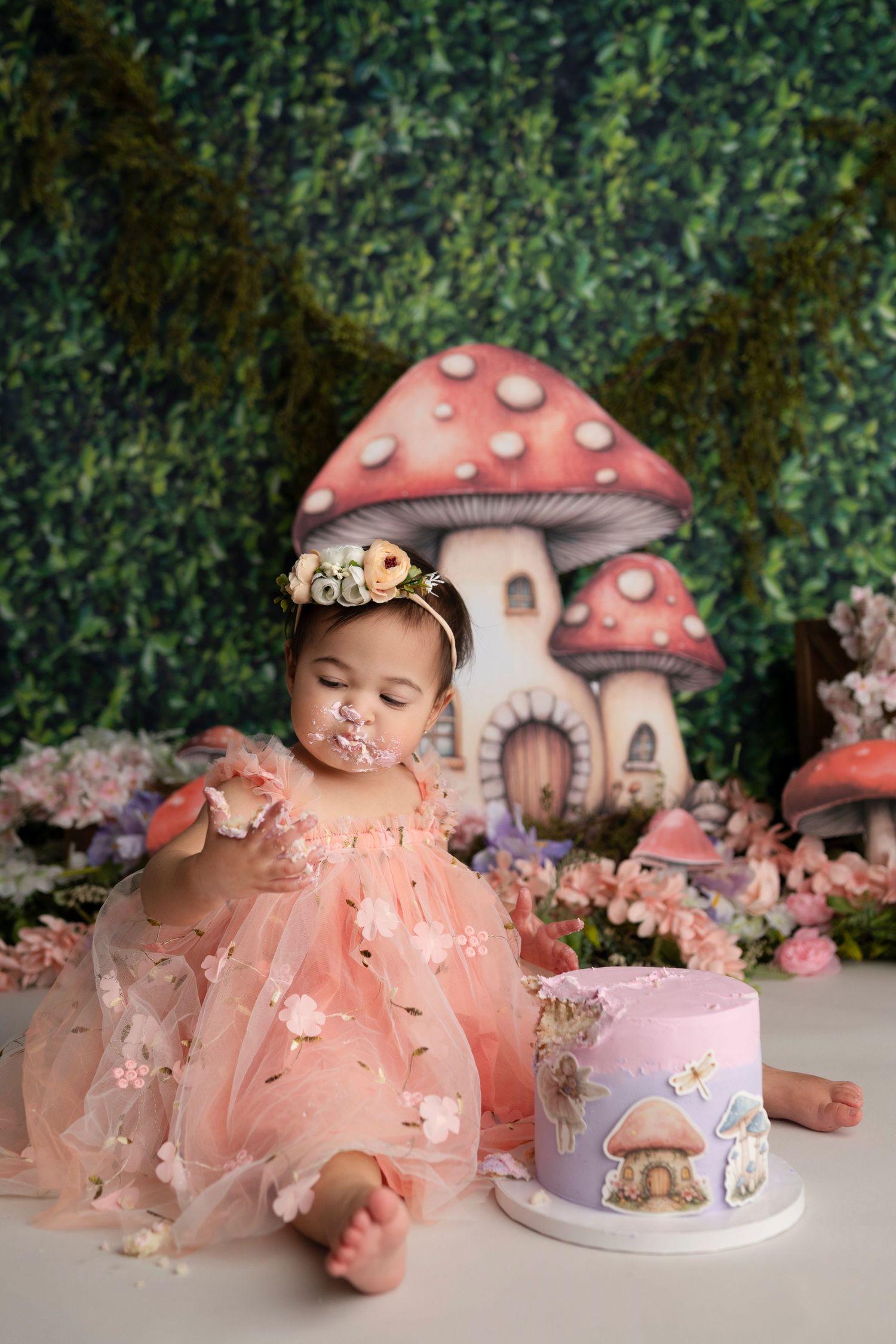 Whimsical fairy garden themed photo setup with pink mushroom houses, flowers, and a decorated cake on display.