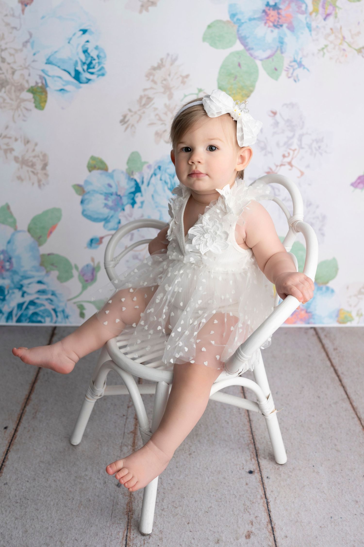 Baby girl in white tulle dress and bow headband sits on white chair against floral backdrop.