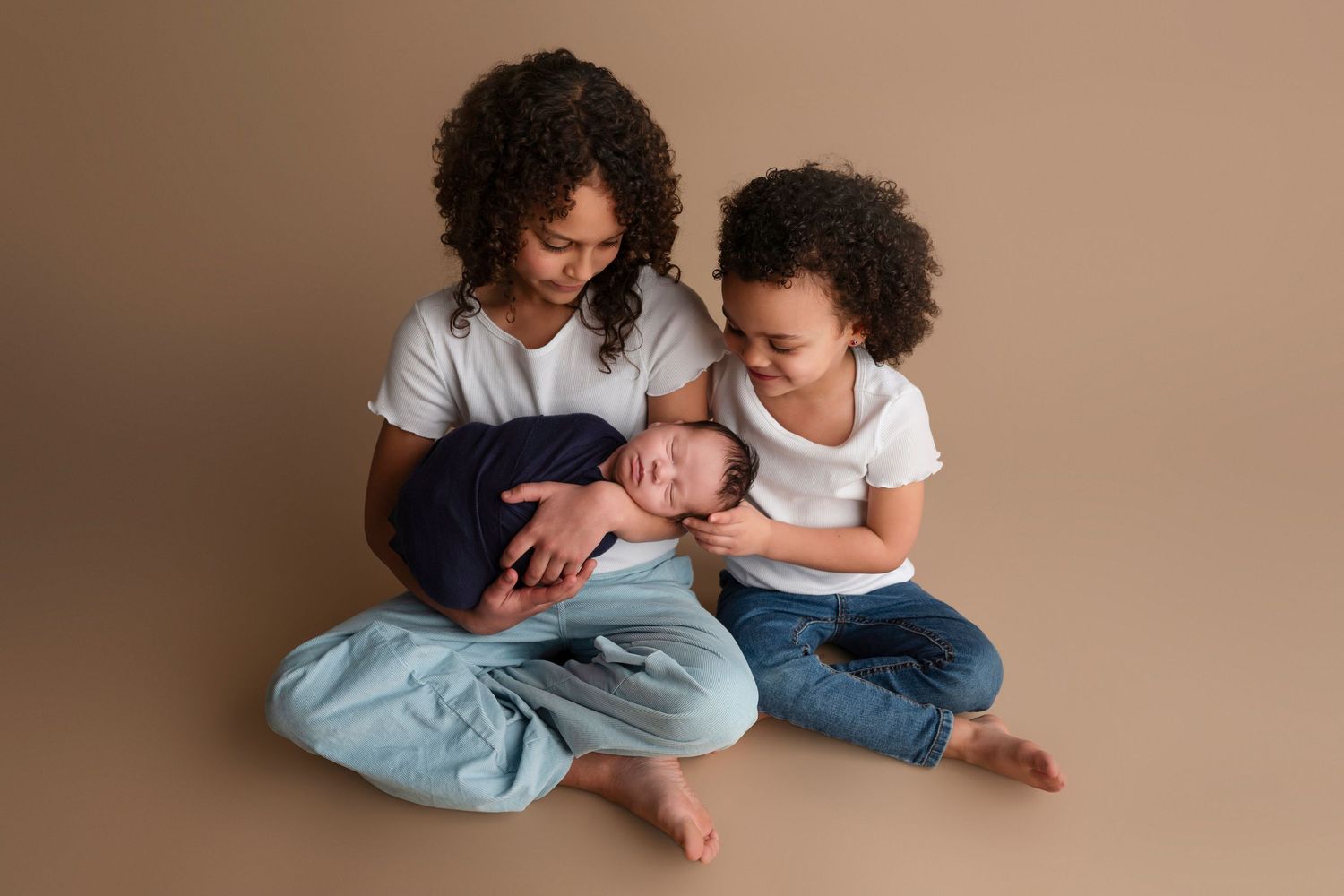 A mother sits cross-legged holding a sleeping newborn in a navy wrap while her toddler leans in lovingly.
