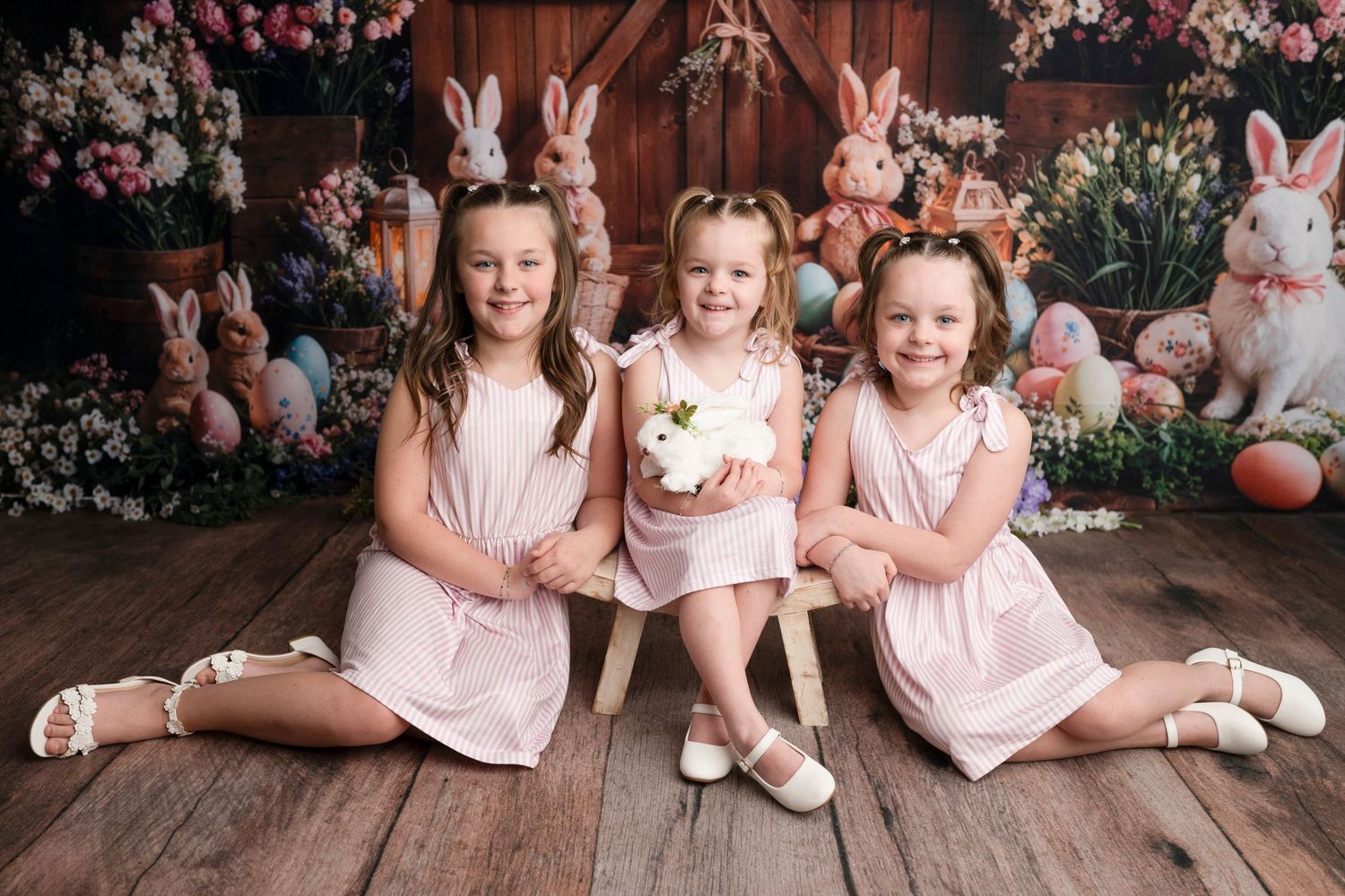Three young girls in matching pink dresses pose with Easter bunnies and colorful eggs in a festive spring photo.