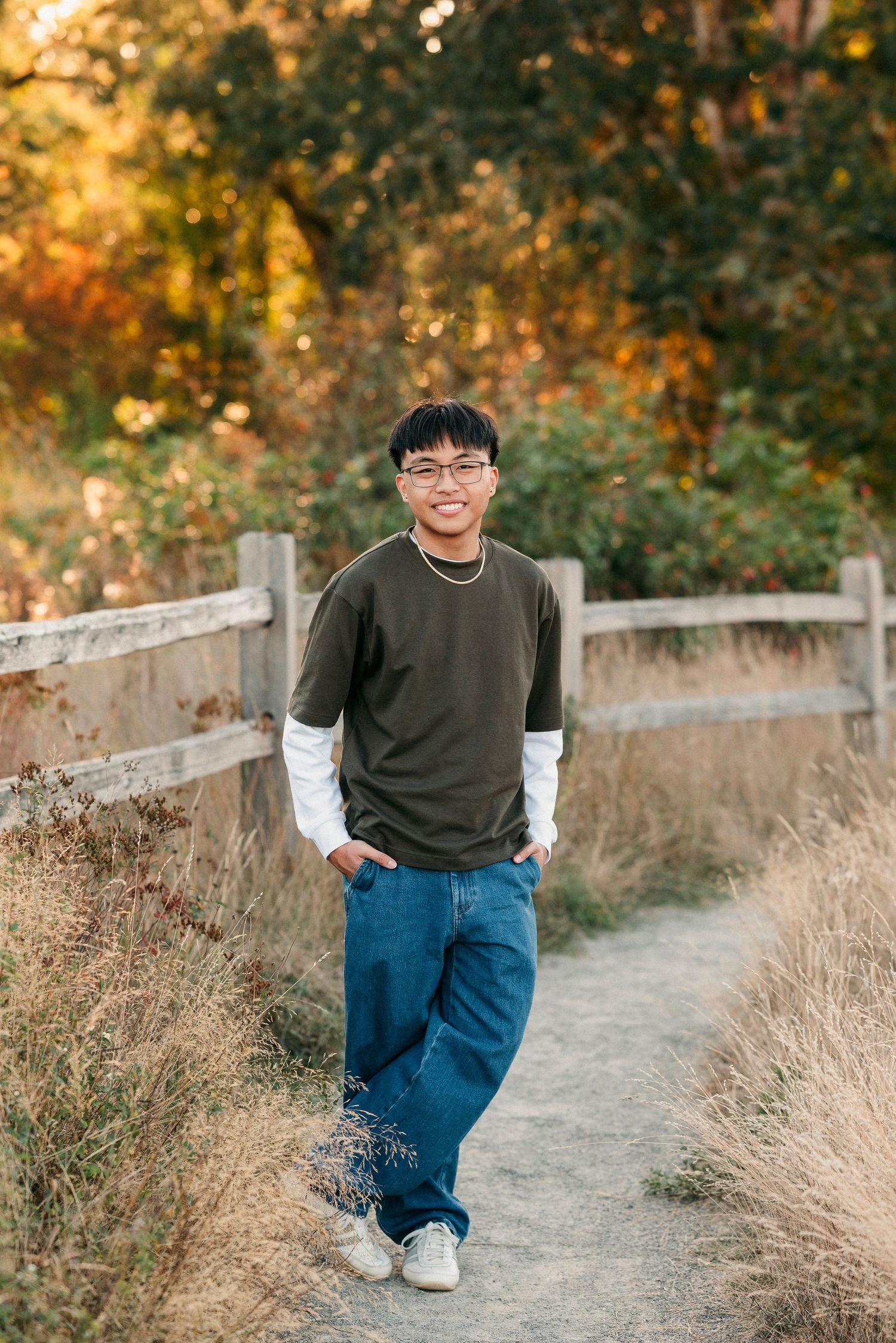 A person in a dark sweater and blue jeans poses casually on a nature trail during golden hour.