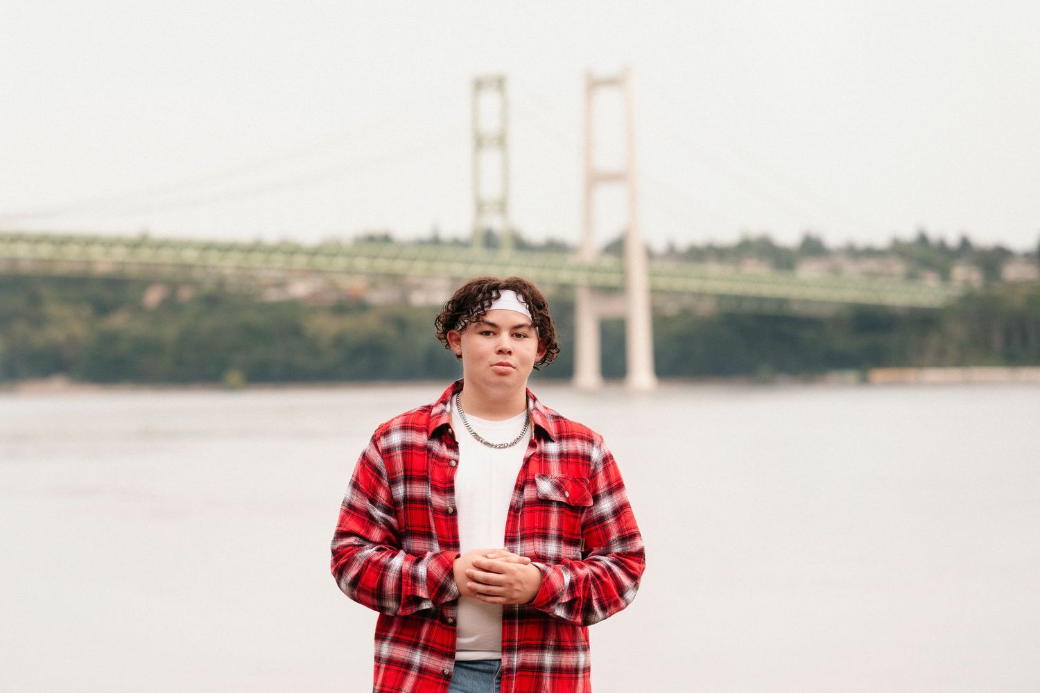 Person in red plaid flannel shirt stands near waterfront with bridge visible in background.