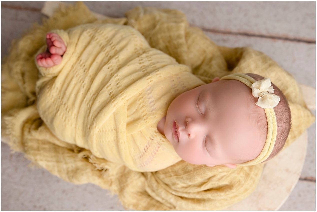 A sleeping newborn wrapped in soft yellow fabric with a delicate floral headband lies peacefully on a wooden surface.