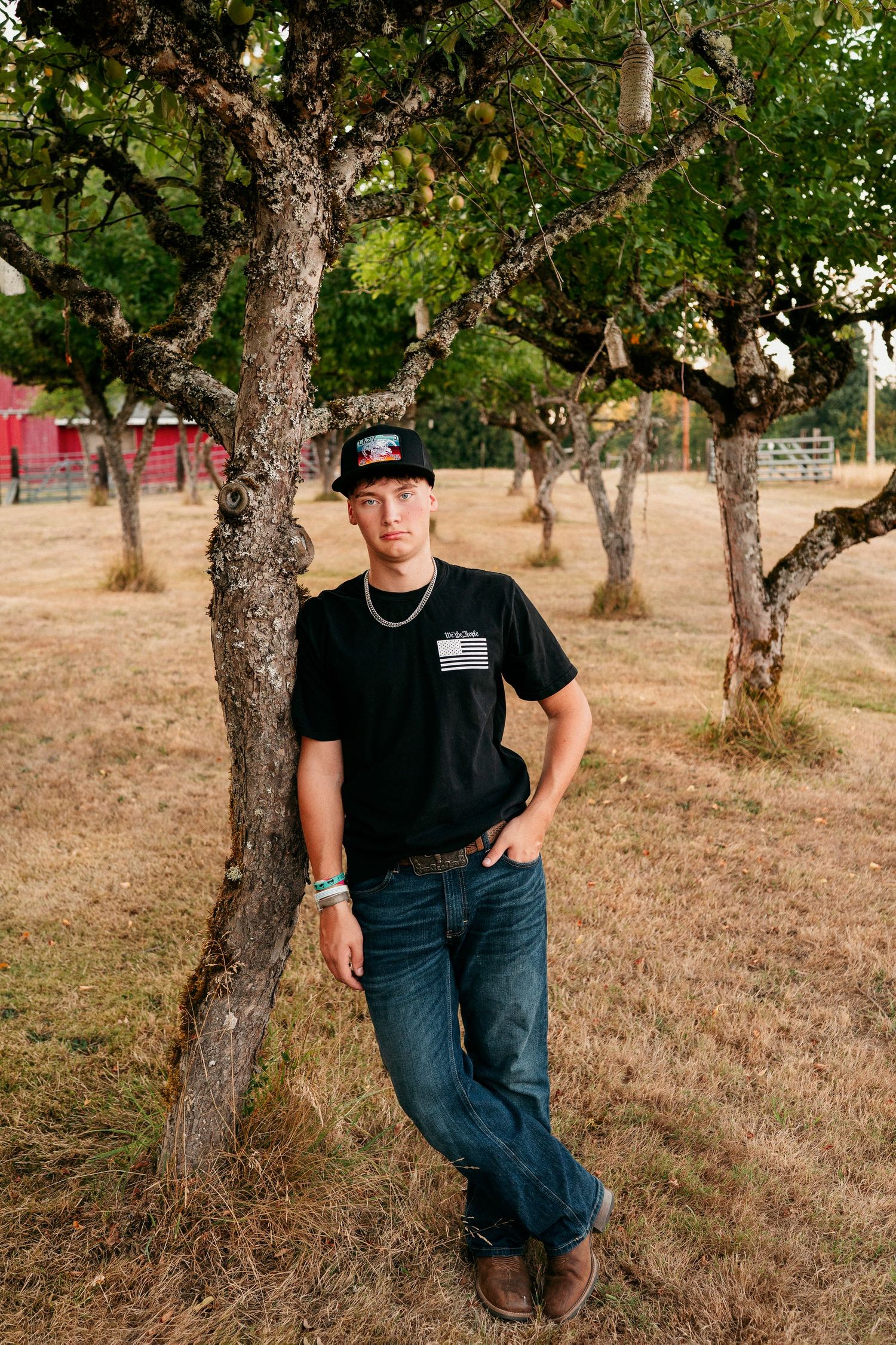 Person in casual black attire and jeans sits on grassy hillside among trees in outdoor setting.