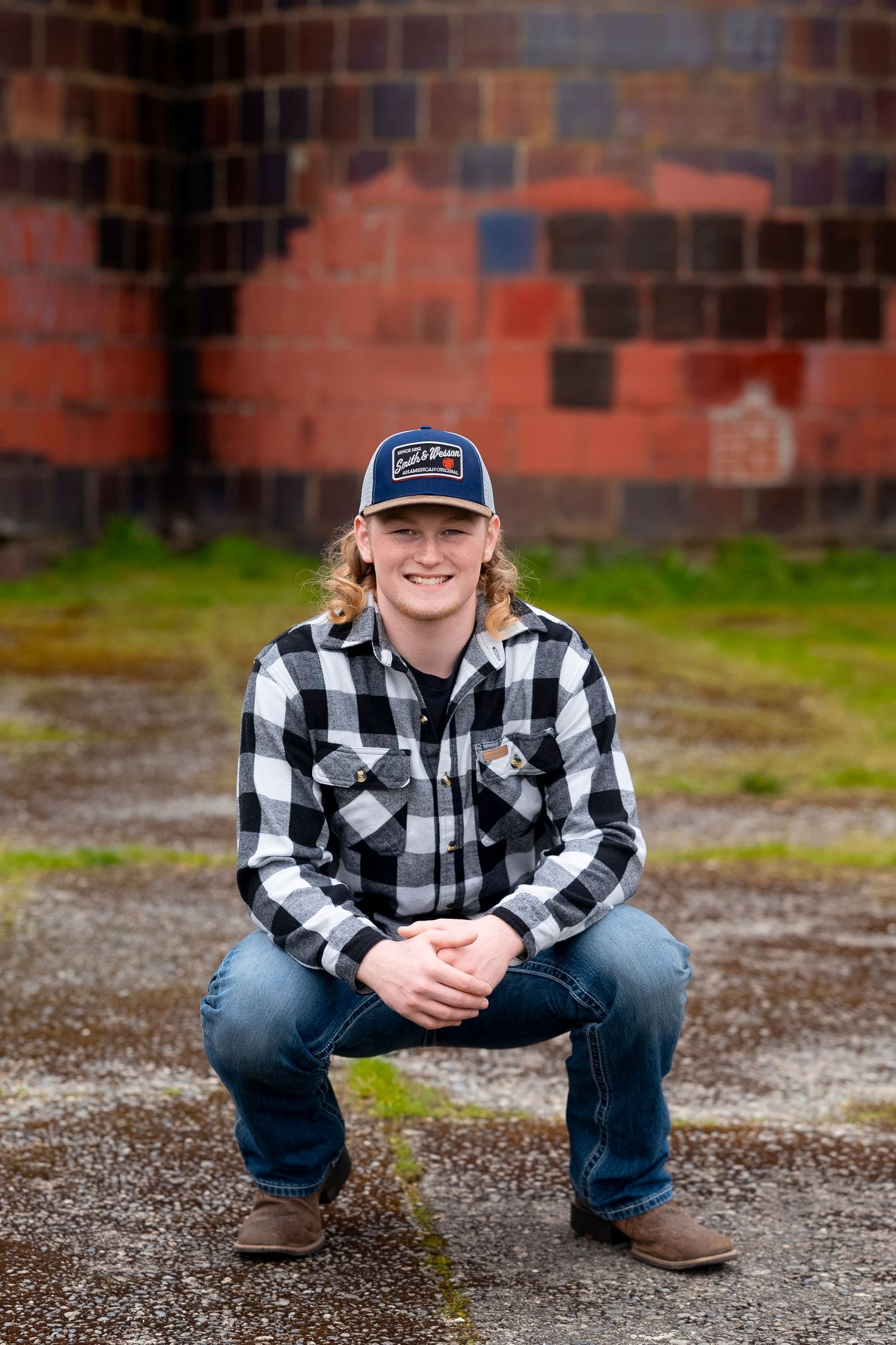 A person wearing a black and white plaid flannel shirt, baseball cap and jeans crouches against a brick wall background.