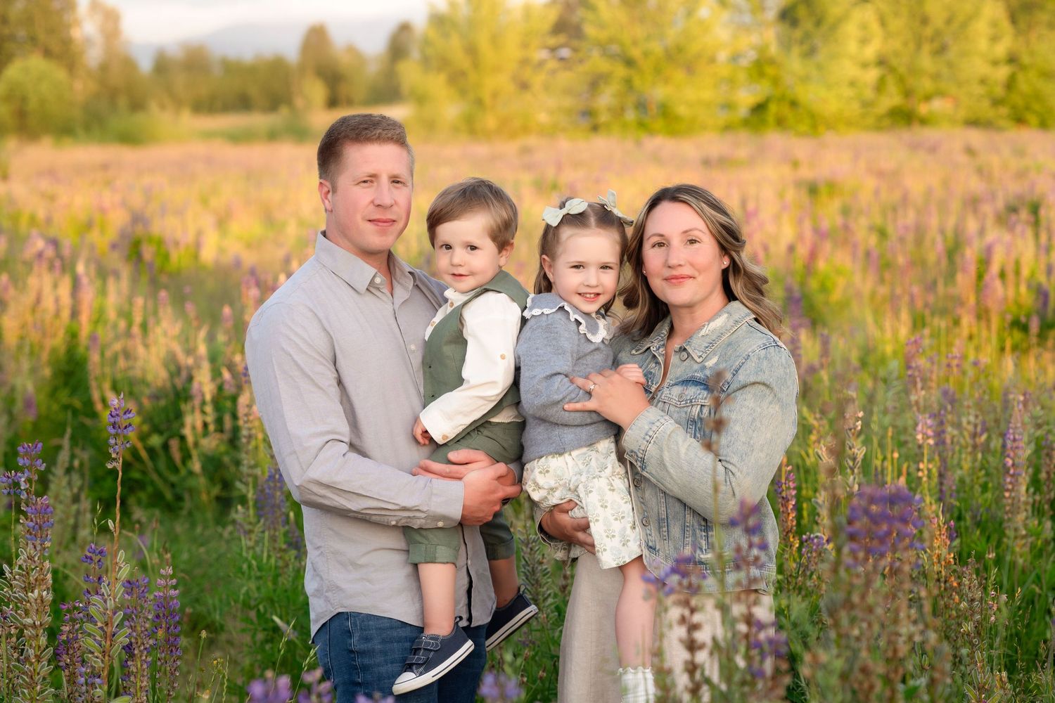 Family of four posing in a colorful wildflower field during golden hour, parents holding two young children.