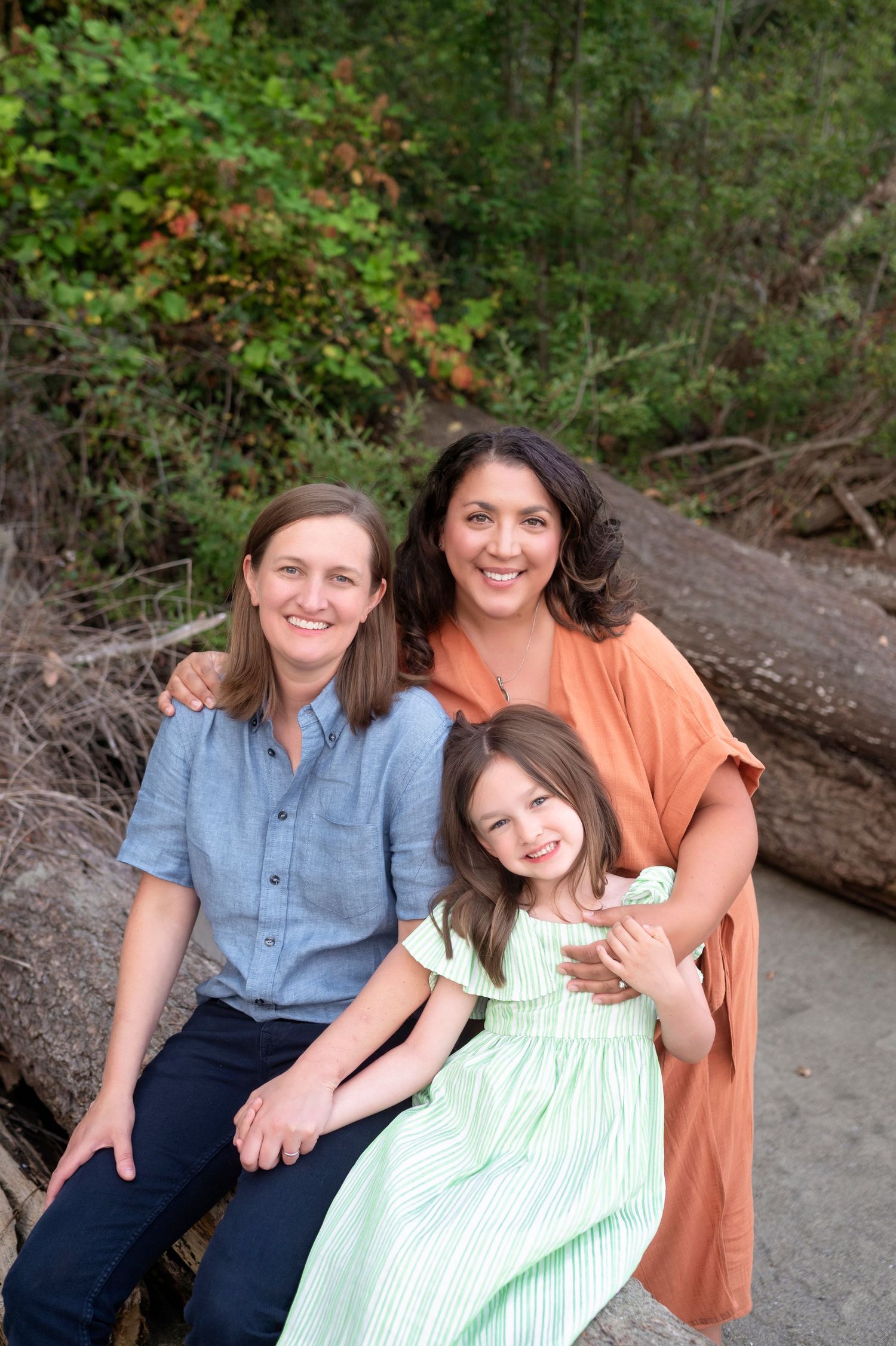Two women and a young girl smile together outdoors, sitting on a log surrounded by lush green trees.
