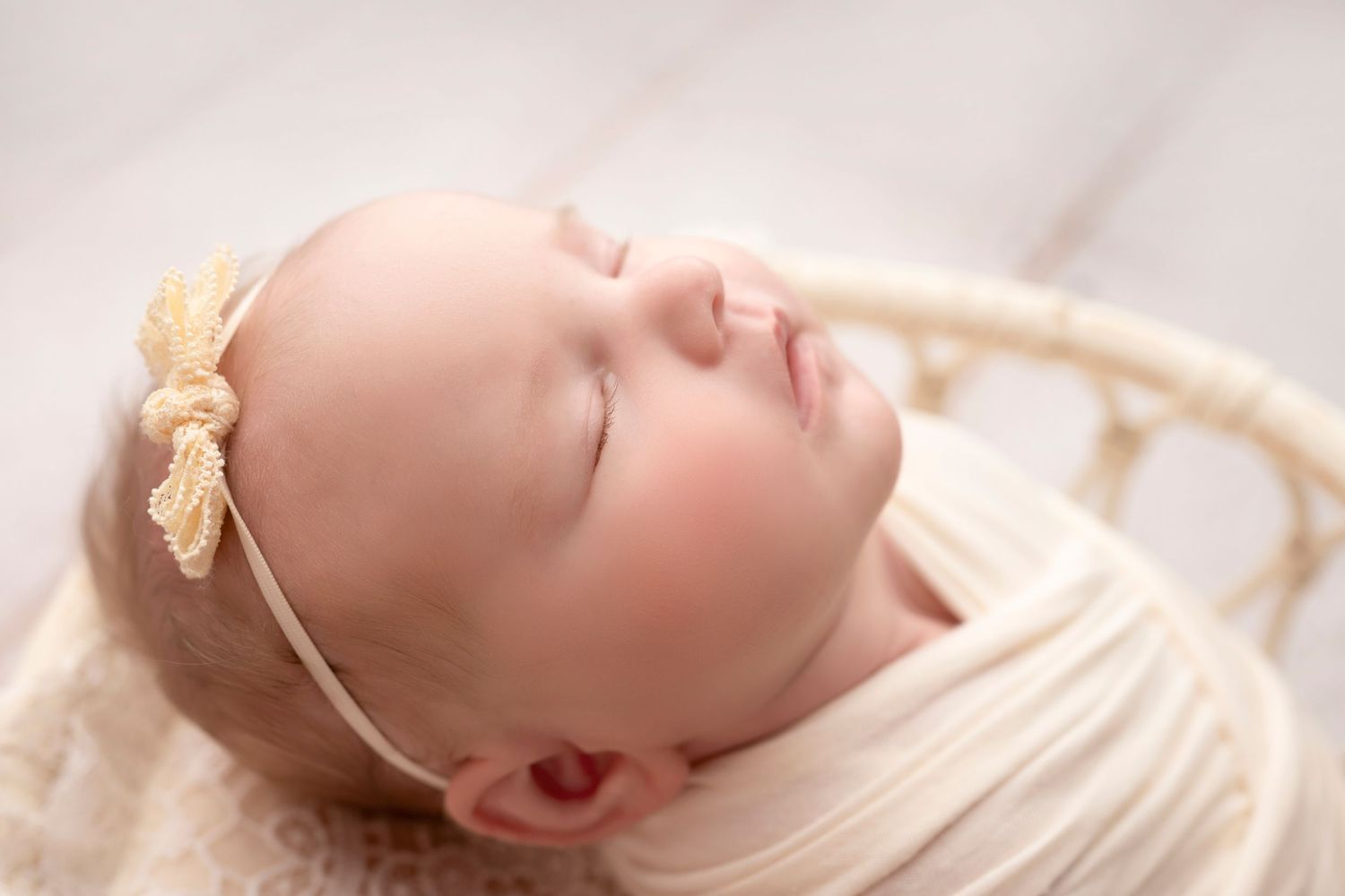 Sleeping newborn baby girl wrapped in cream swaddle with gold bow headband in a white wicker basket.