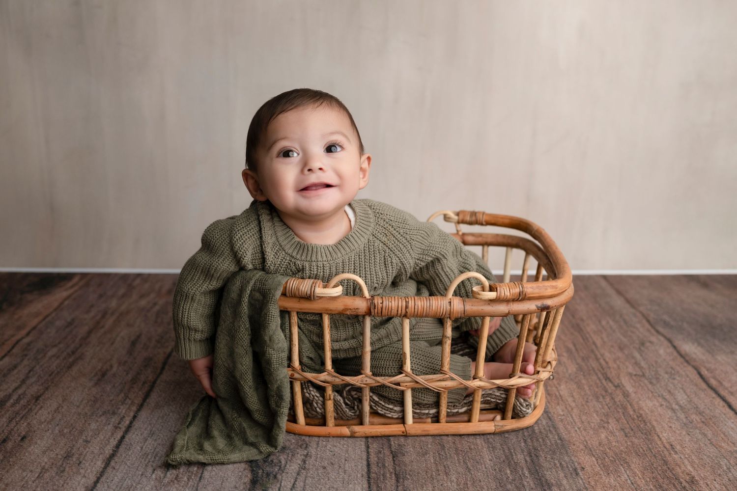 Smiling baby in green knit sweater sitting in a wicker basket on a wood floor with a neutral backdrop.
