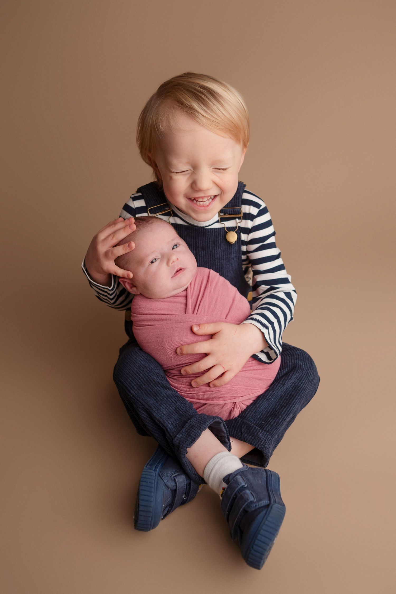 A sibling in a striped shirt tenderly holds and interacts with a sleeping newborn baby on a beige backdrop.