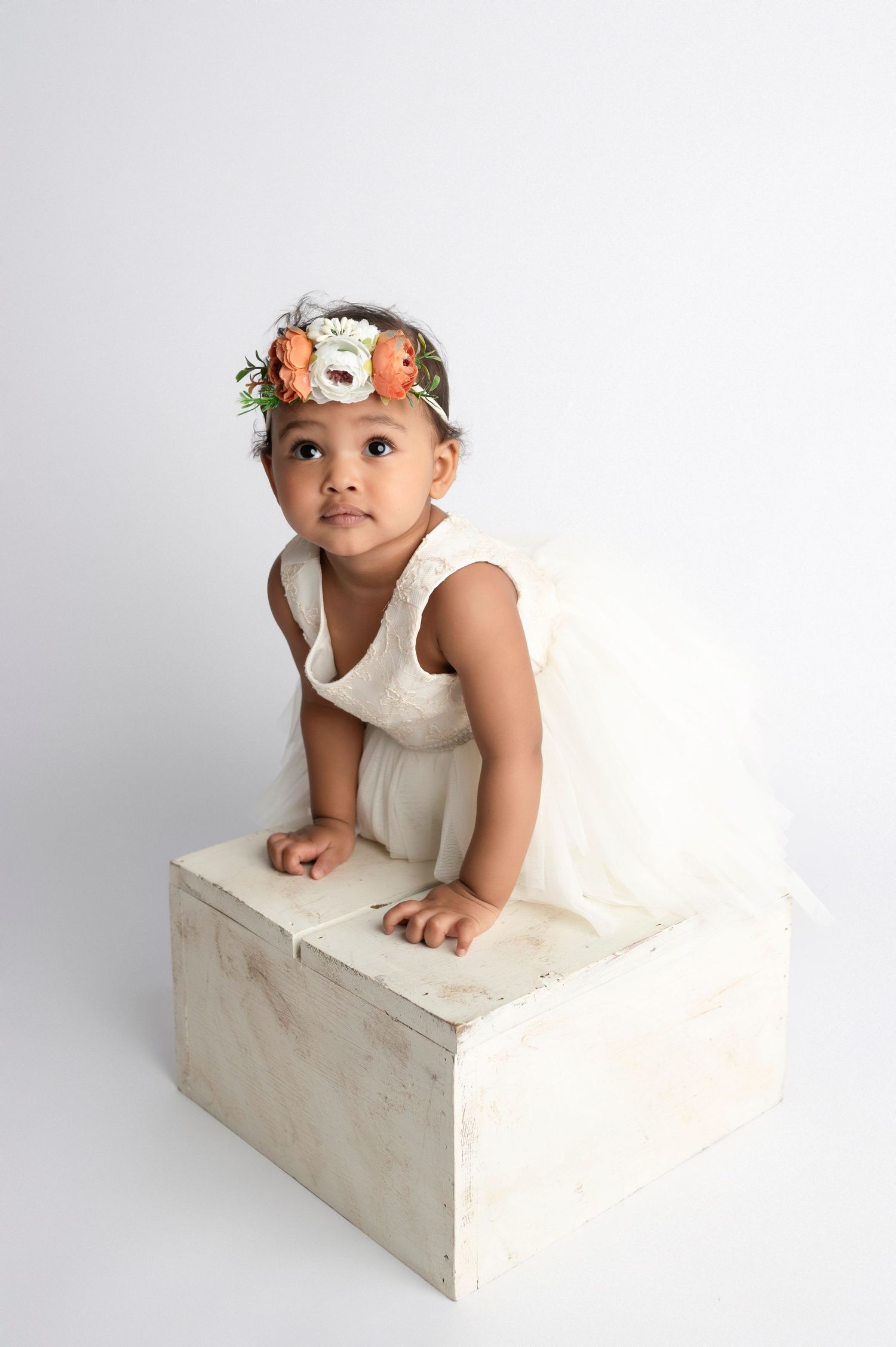 Toddler girl in white dress and floral crown poses on a white wooden box against a light background.