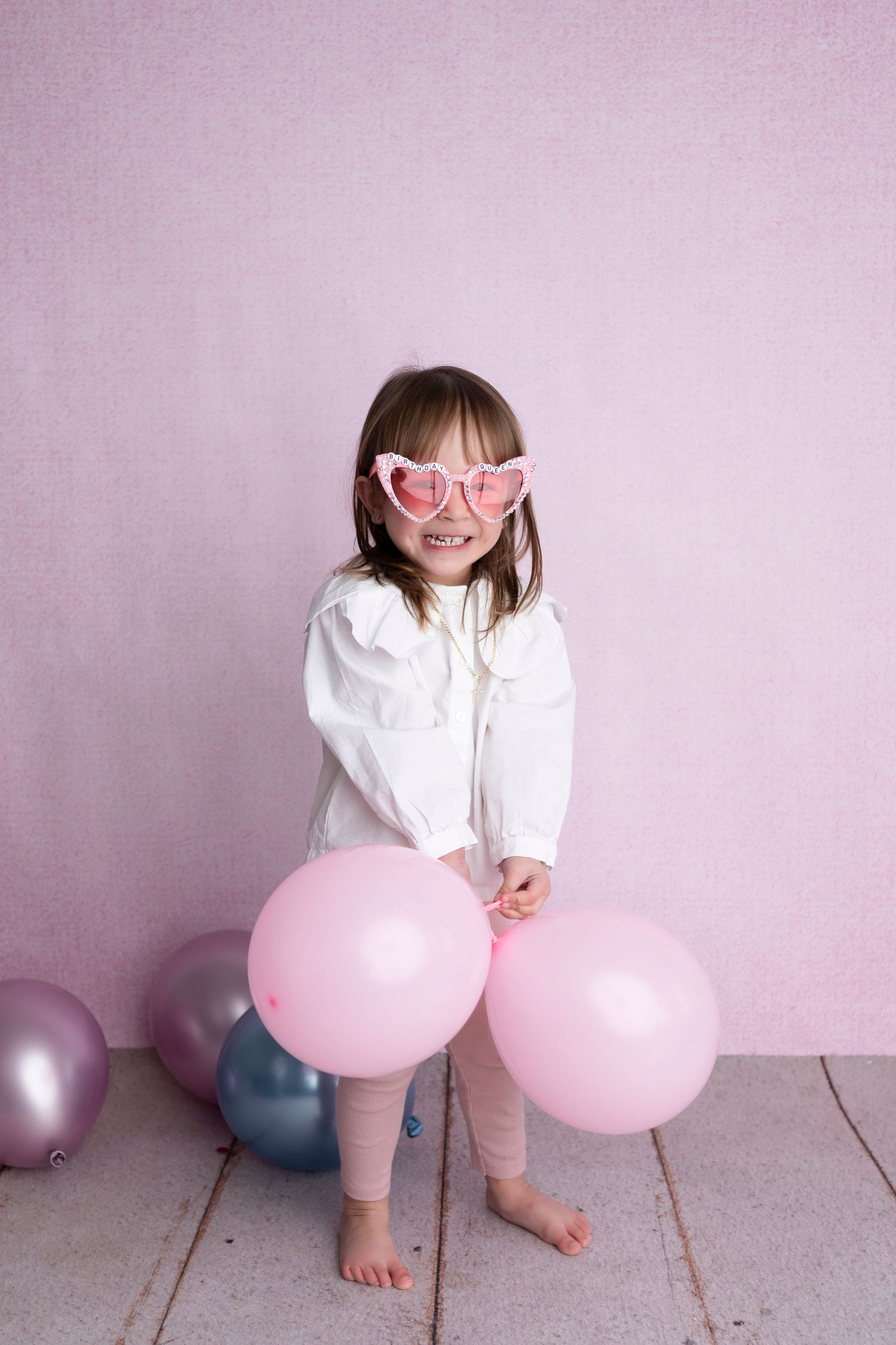 Young girl in white jacket and pink heart sunglasses smiling with pink and silver balloons on pink background.