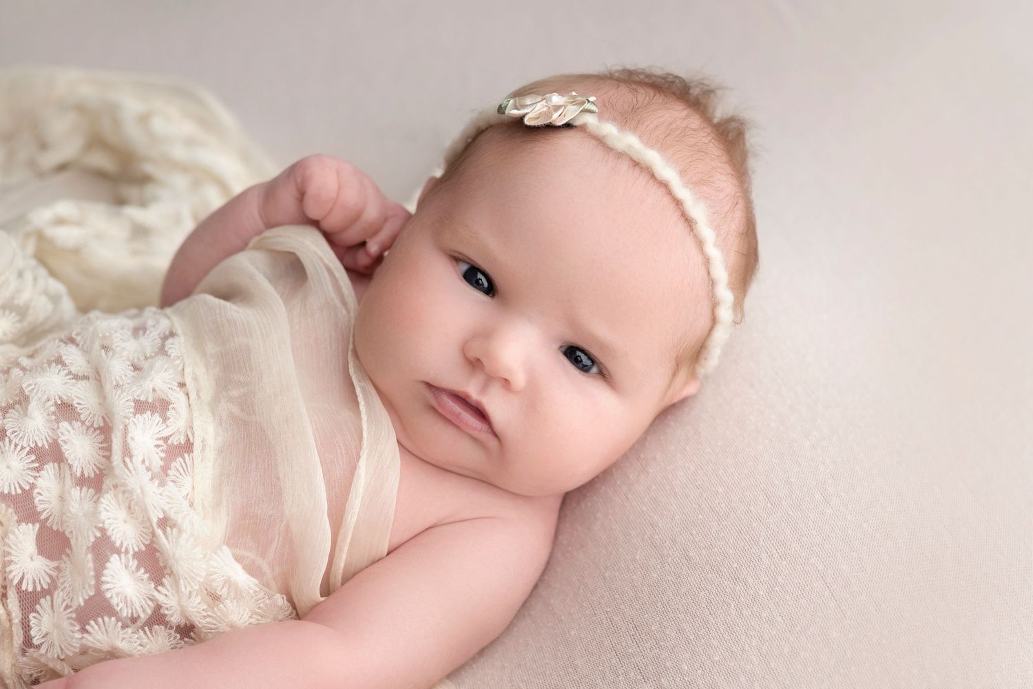 A serene newborn baby girl in a cream lace wrap and floral headband lies on a soft white backdrop.