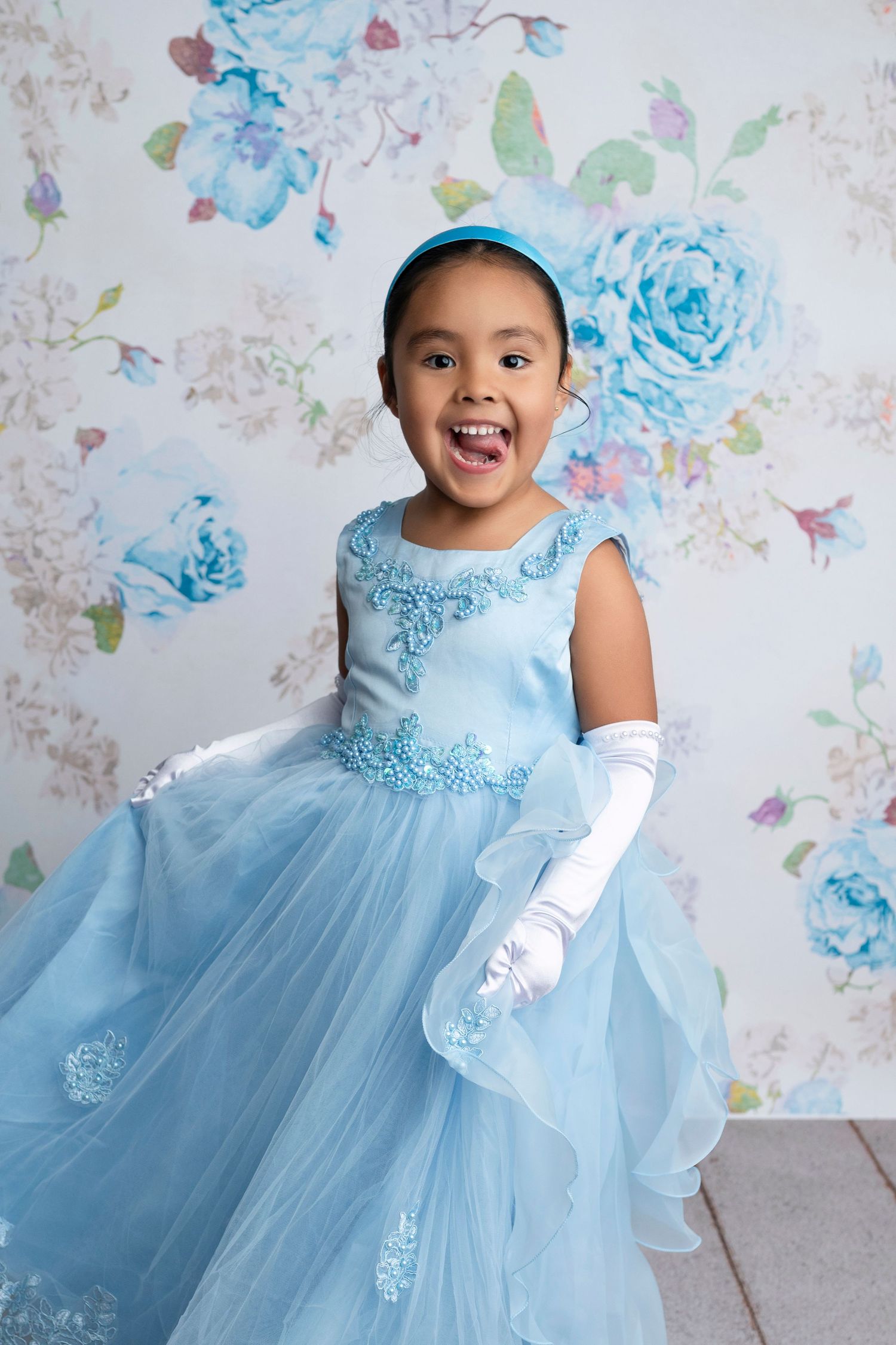 Joyful young girl in a blue princess gown with white gloves and headband, posing before a floral backdrop.