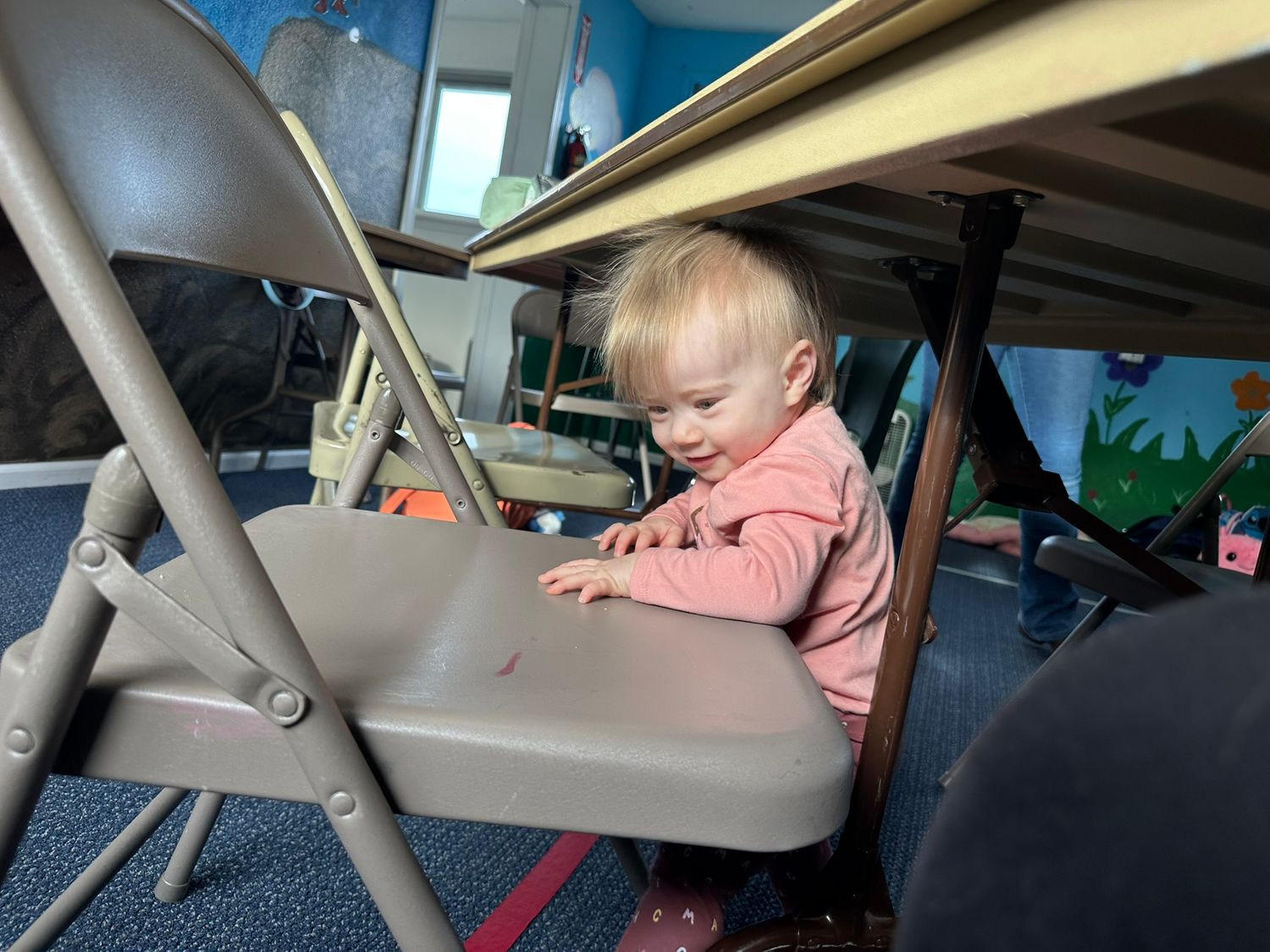 Toddler in pink shirt playing under a table on a folding chair in a colorful classroom setting.