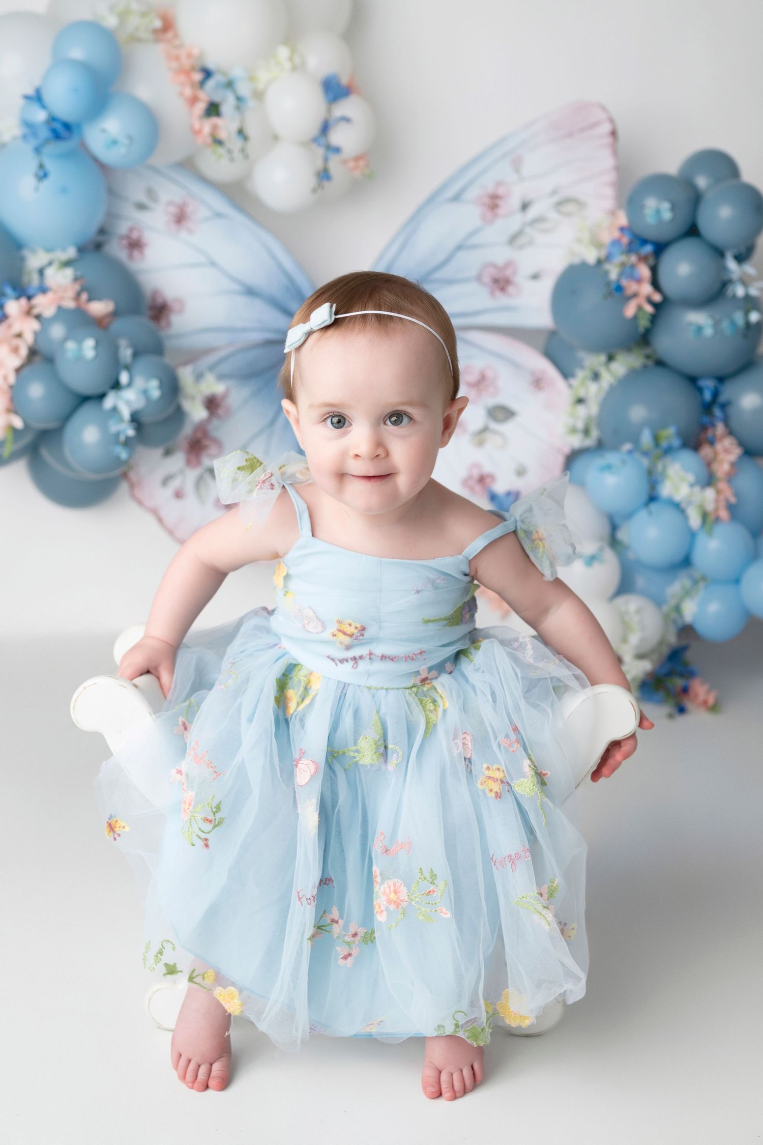 Baby in light blue tulle dress with floral details poses against backdrop of blue balloons and butterfly decorations.
