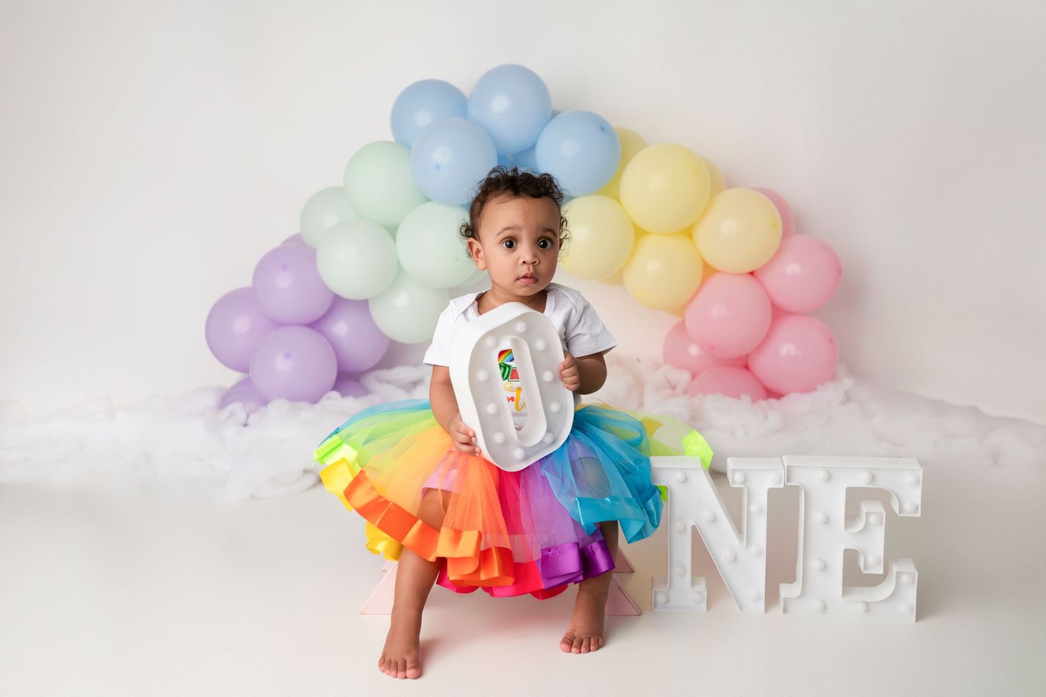 A toddler in rainbow tutu and white shirt poses under pastel balloon arch with 'ONE' letters.