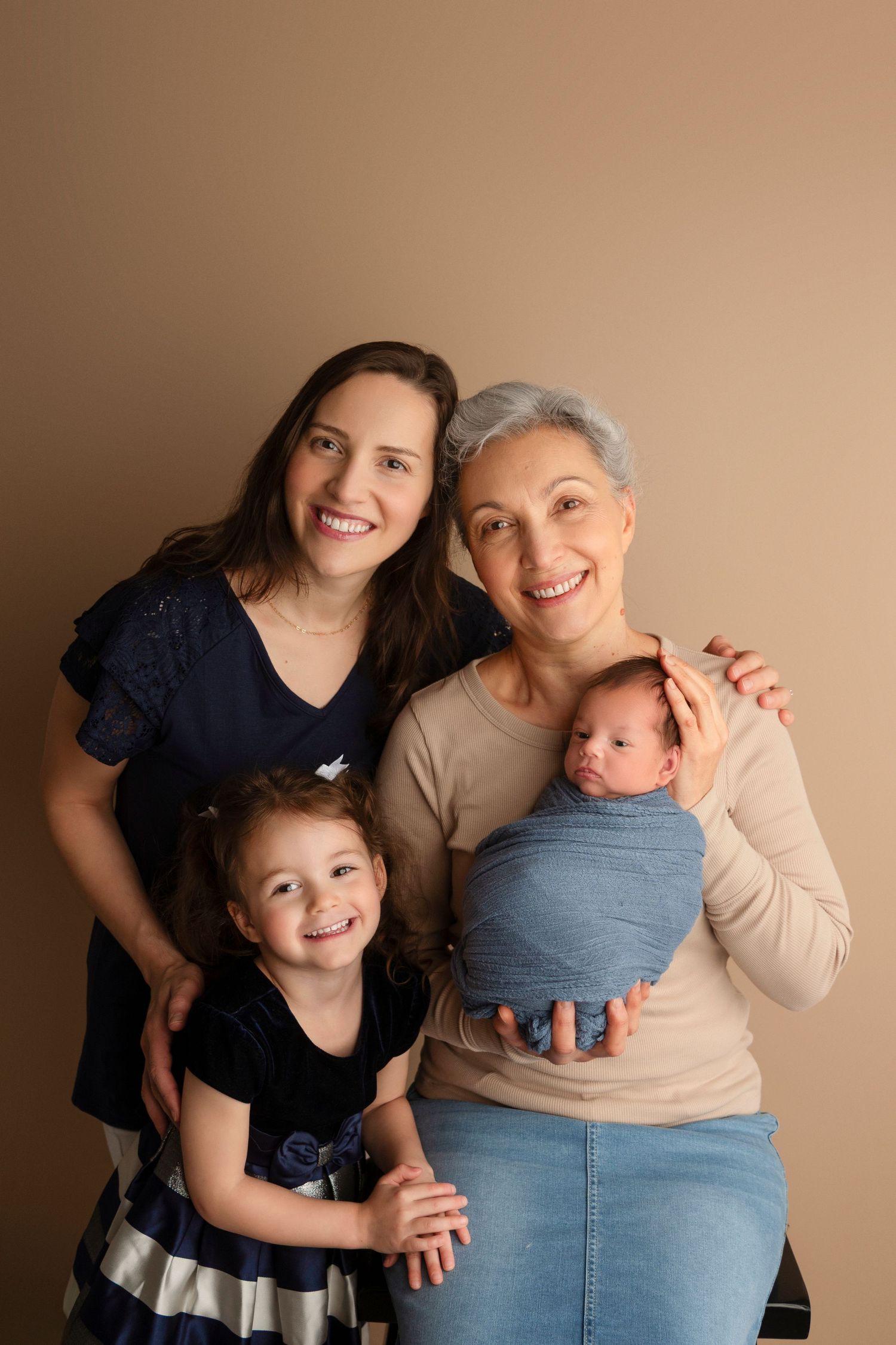 Three generations pose together for a heartwarming family portrait against a neutral brown background.