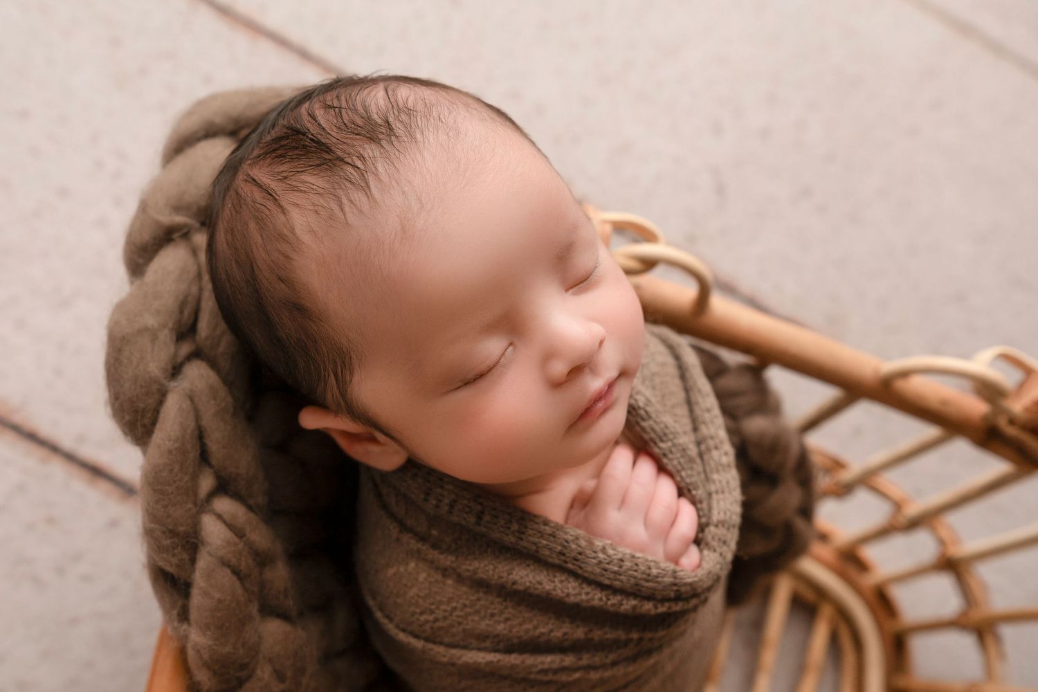 Newborn baby sleeping peacefully in brown swaddle blanket during professional photography session.
