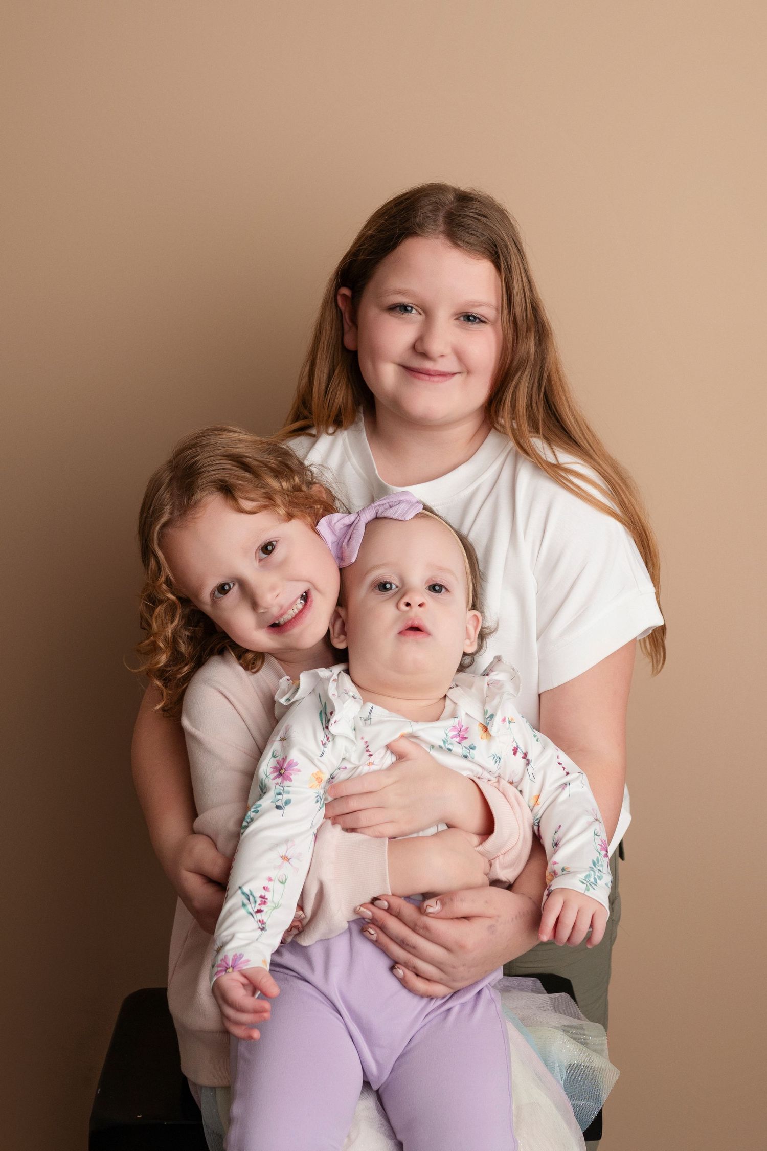 Three sisters pose together, oldest girl holding toddler and baby in a studio portrait with beige background.