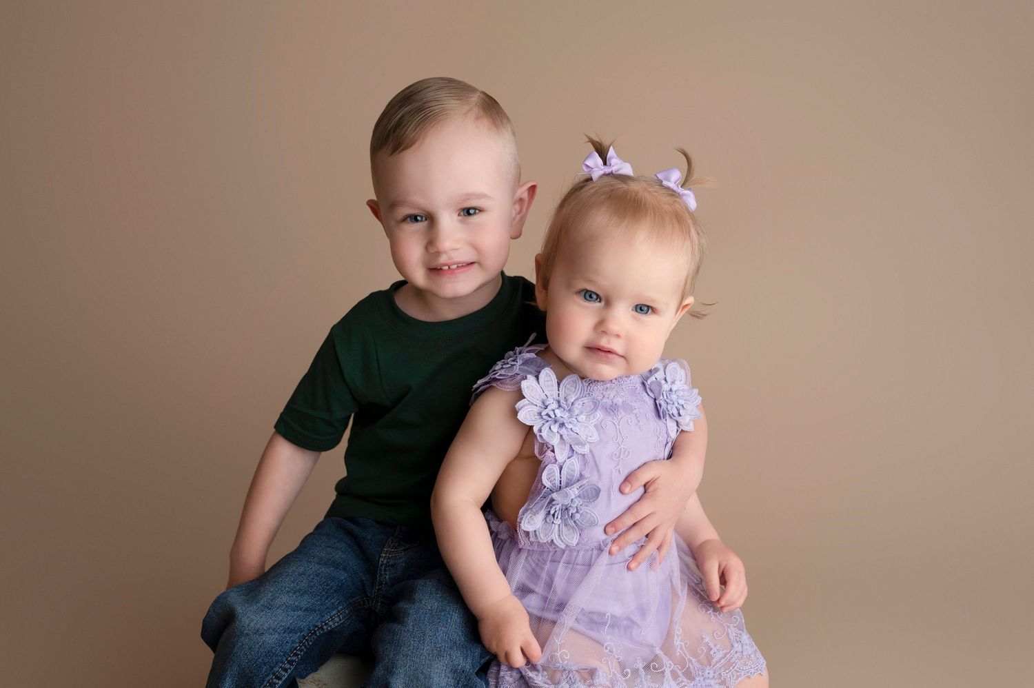 A young boy in a green shirt sits with a baby girl in a purple floral dress and bow headband for a studio portrait.