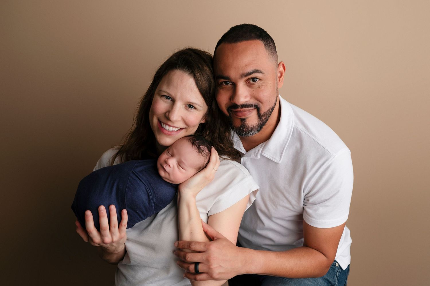 A couple smiles warmly while cradling their swaddled newborn baby in a professional studio portrait.