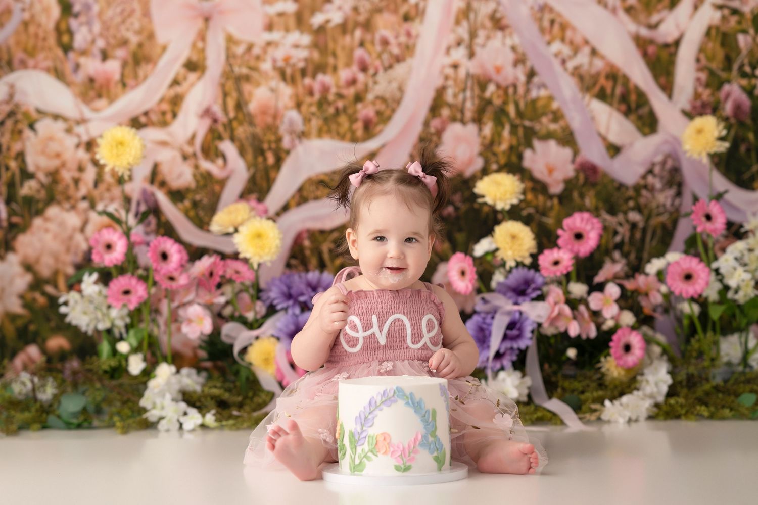 Baby girl in pink tutu celebrates first birthday with floral backdrop and cake smash setup.