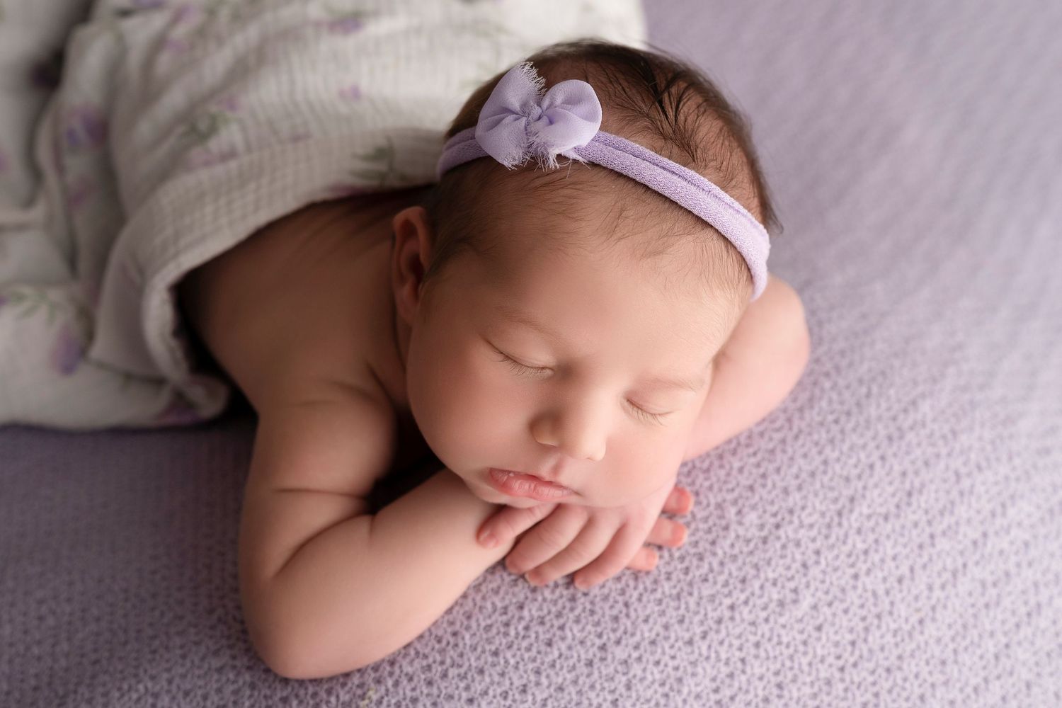 Sleeping newborn baby girl wearing a purple floral headband, resting on a lavender knit blanket.
