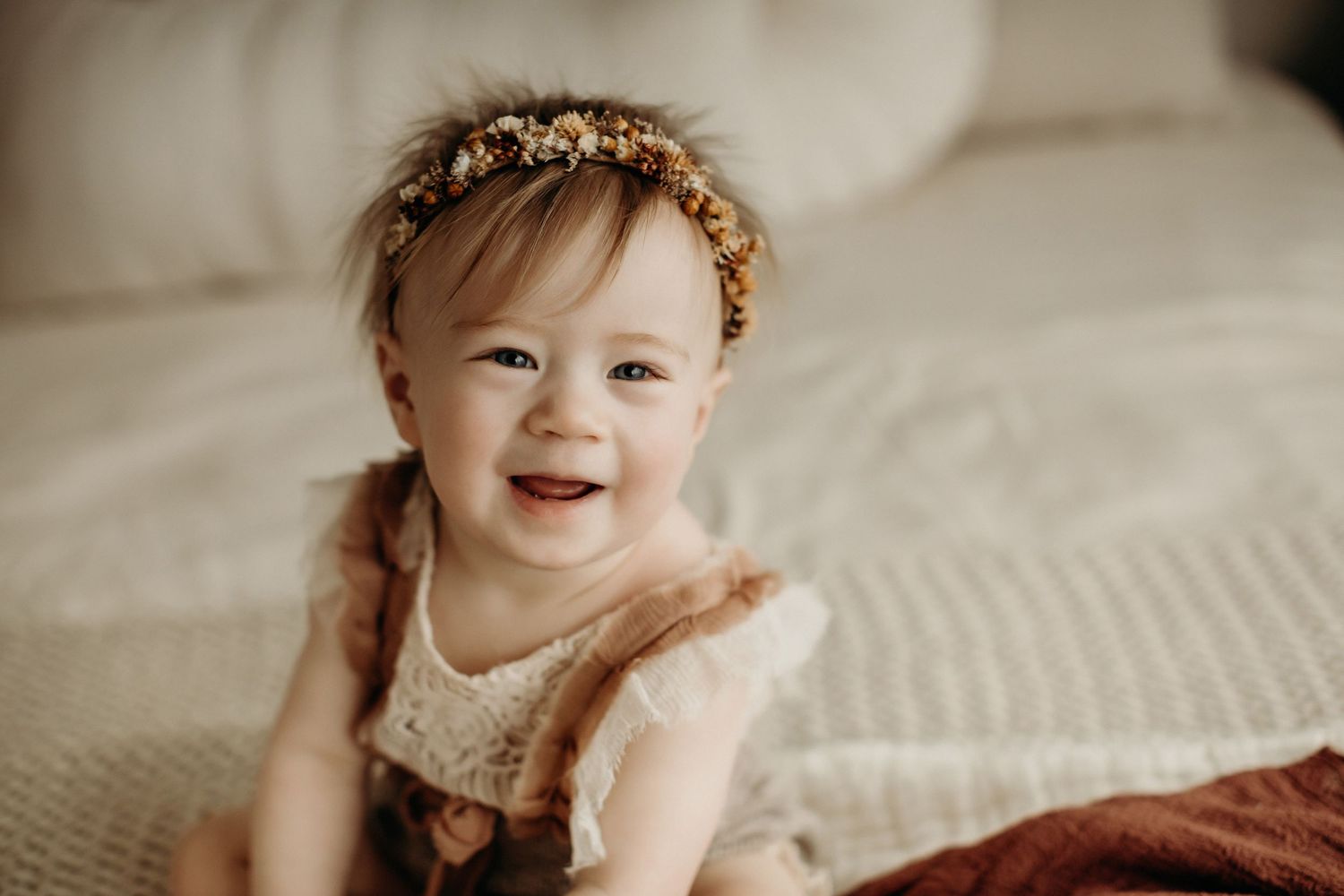 A smiling baby wearing a cream lace dress sits on a textured cream blanket with a delicate floral headband.