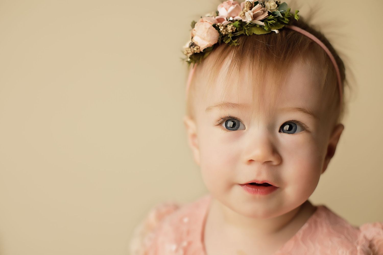 Baby wearing a pink dress and floral headband smiles at camera against beige background.
