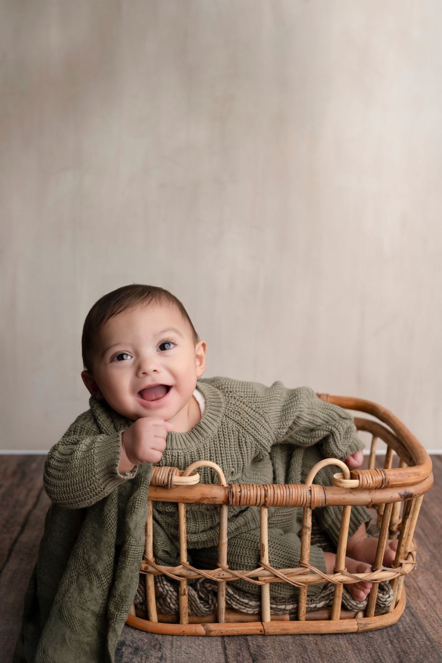 Smiling baby in green knit sweater sitting in a wicker basket on a wooden floor with neutral backdrop.