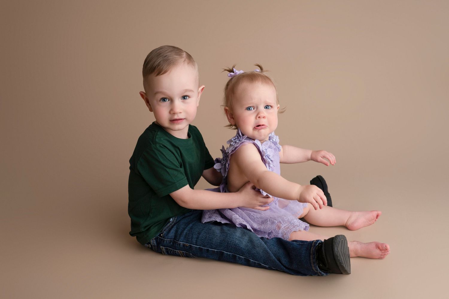 Toddler boy in green shirt with baby sister in purple romper sitting together on beige backdrop.