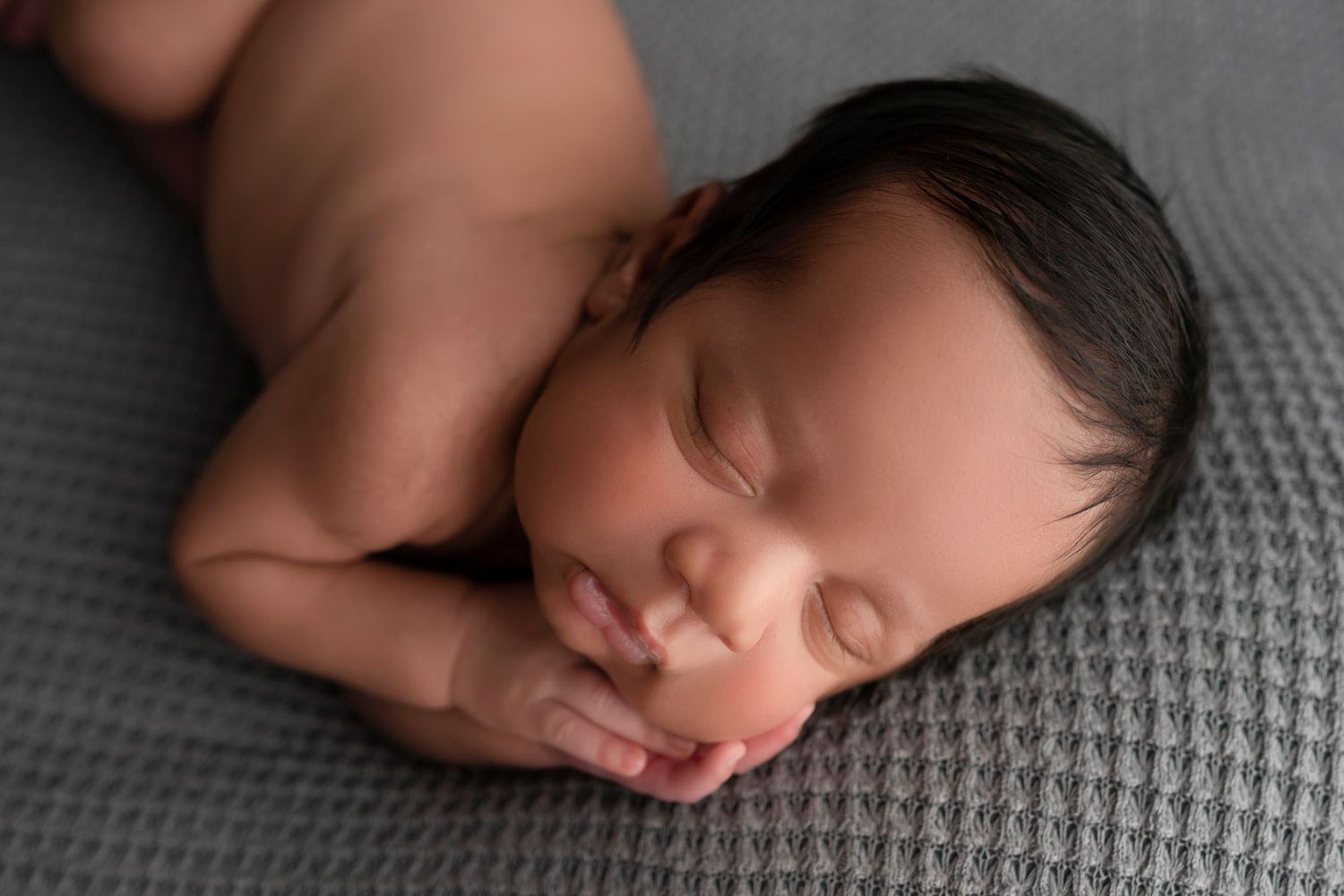 Peaceful sleeping newborn baby with dark hair resting on a gray textured blanket in a professional portrait.