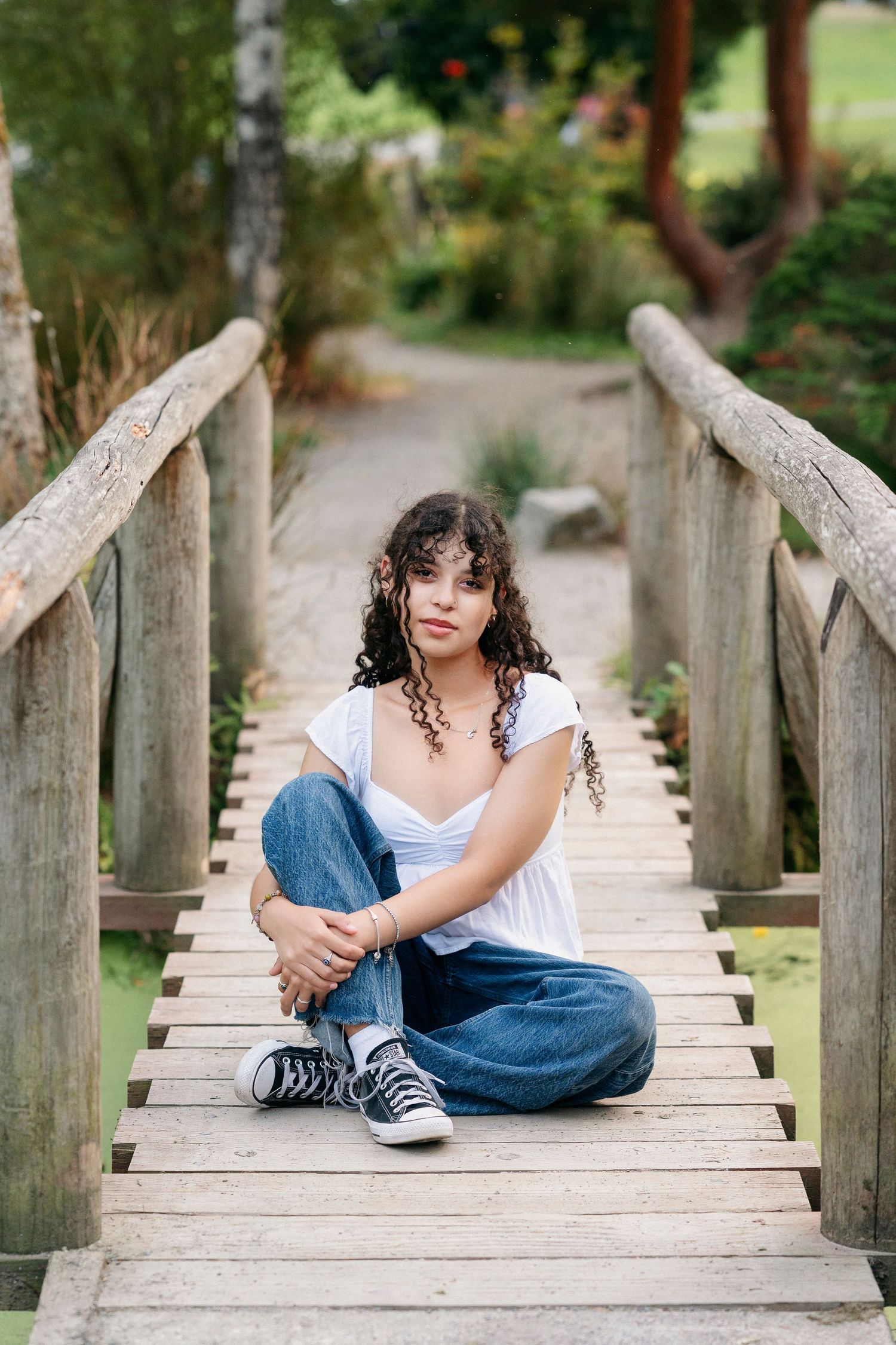 A casual portrait on a wooden footbridge in a lush garden setting, featuring jeans, sneakers and a white top.