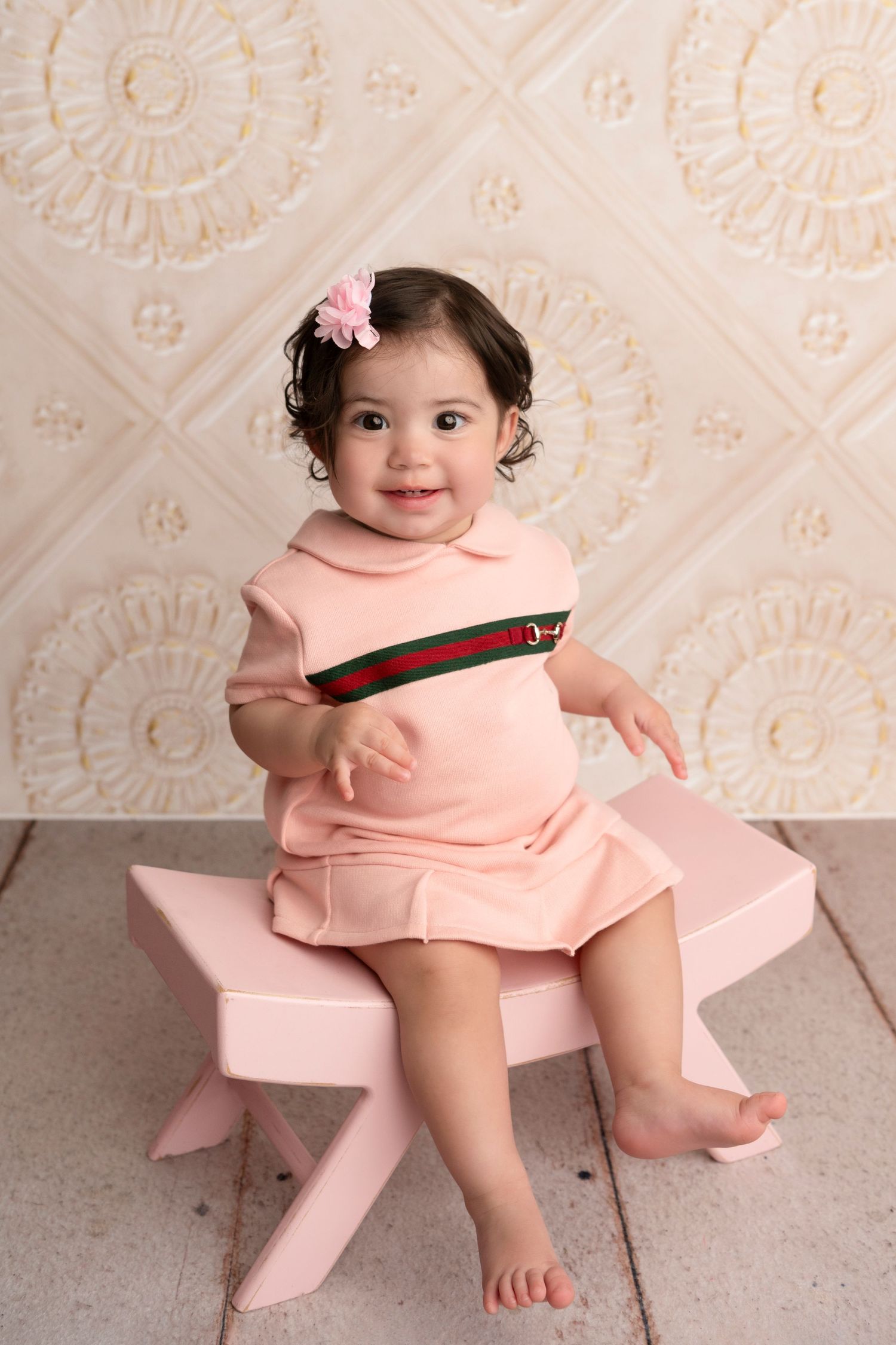 A small child in a pink dress sits on a pink stool against a floral wallpaper background.