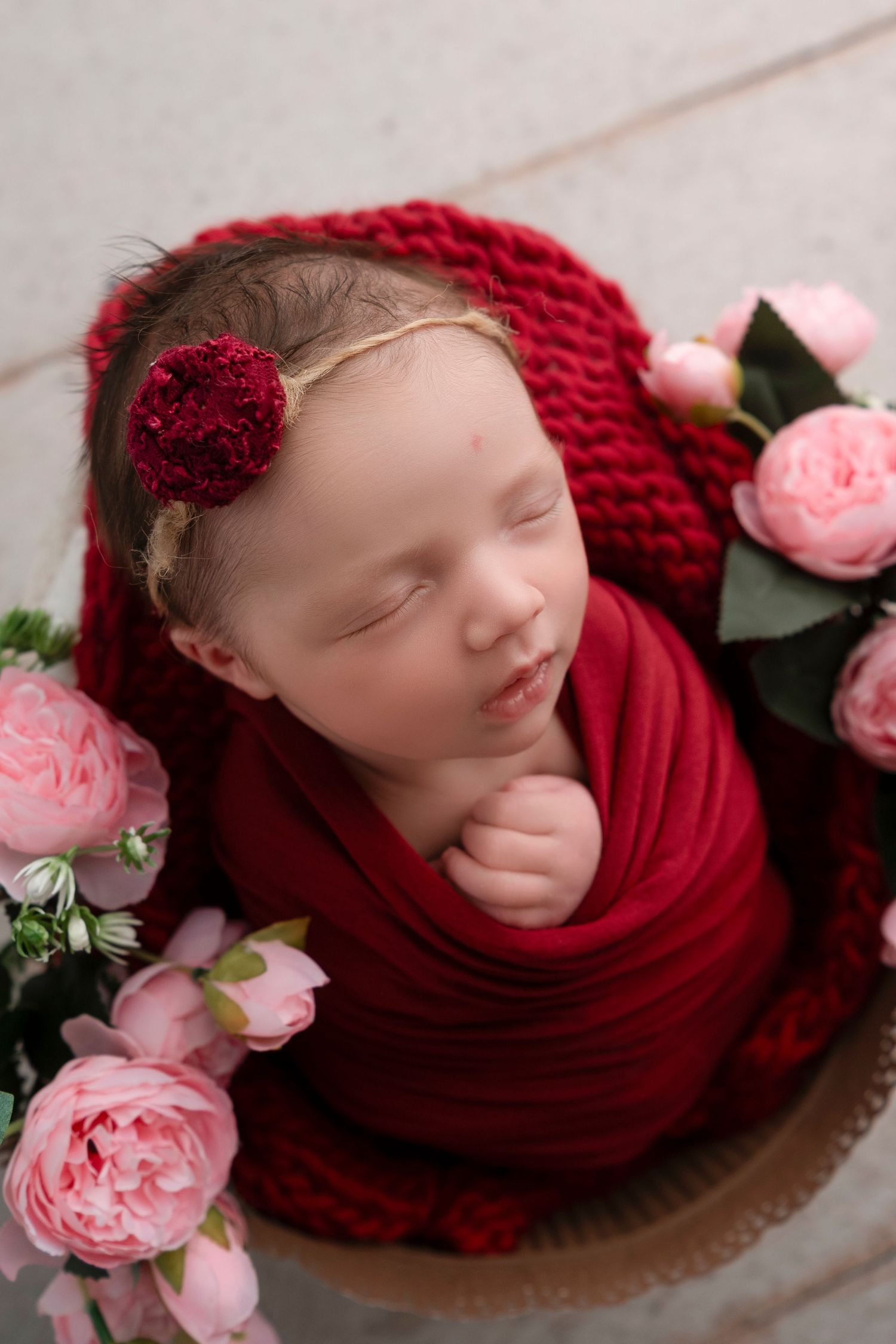Newborn photography with red knit wrap and delicate pink roses creating a romantic and peaceful setting.