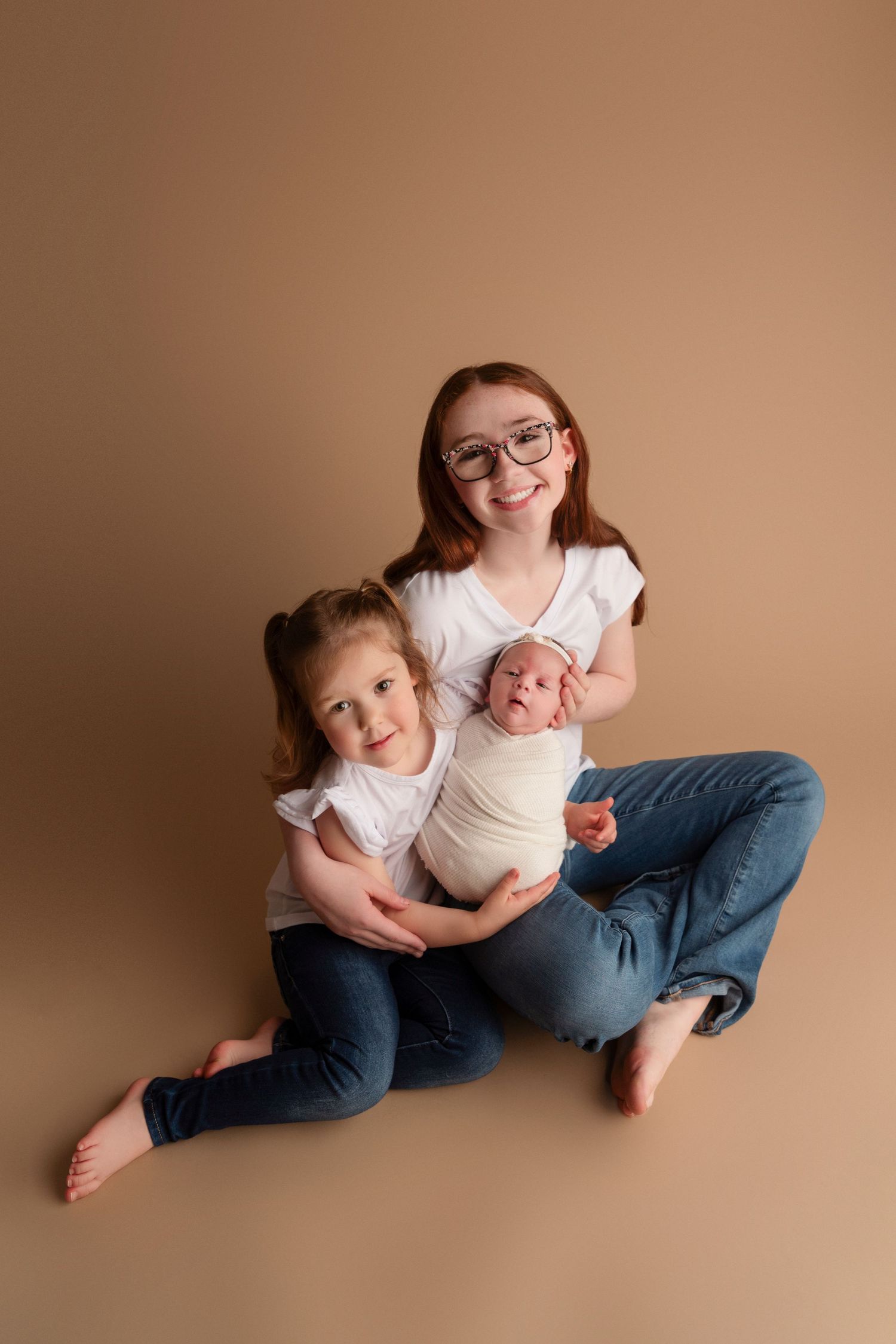 Three siblings sit together on a beige background wearing white tops and jeans in a casual studio portrait.