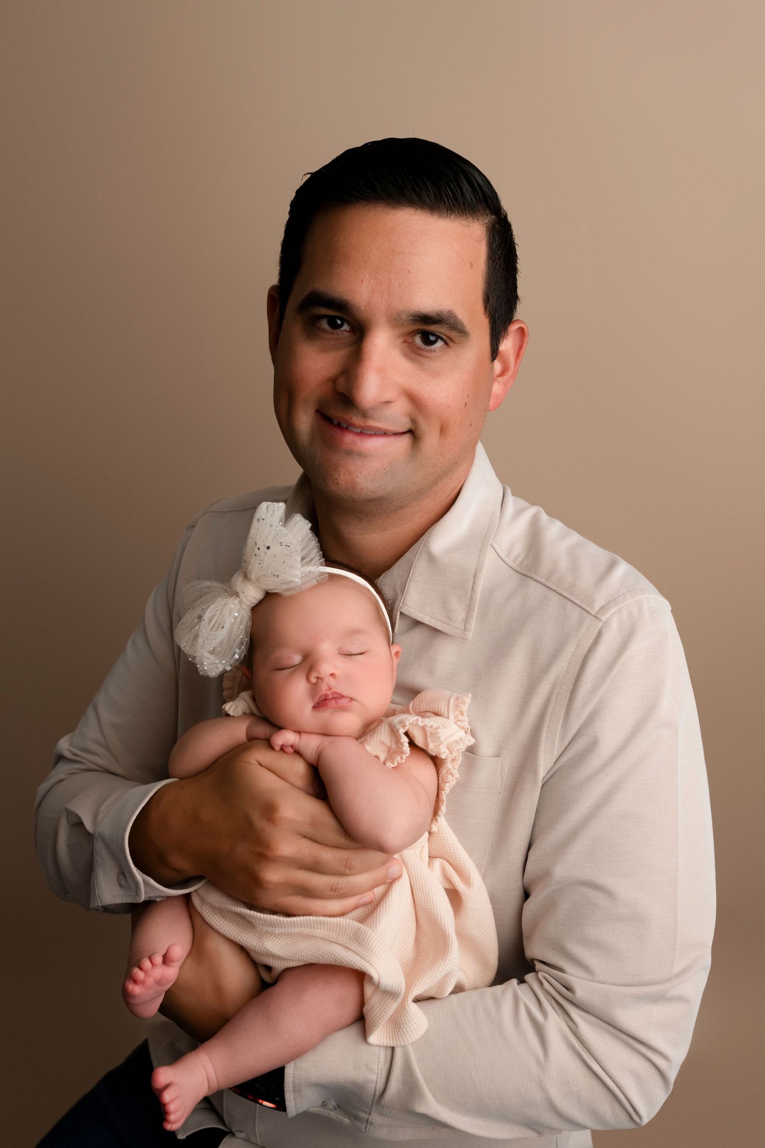 A tender portrait of a parent holding a sleeping newborn baby wearing a peach-colored outfit against a neutral background.