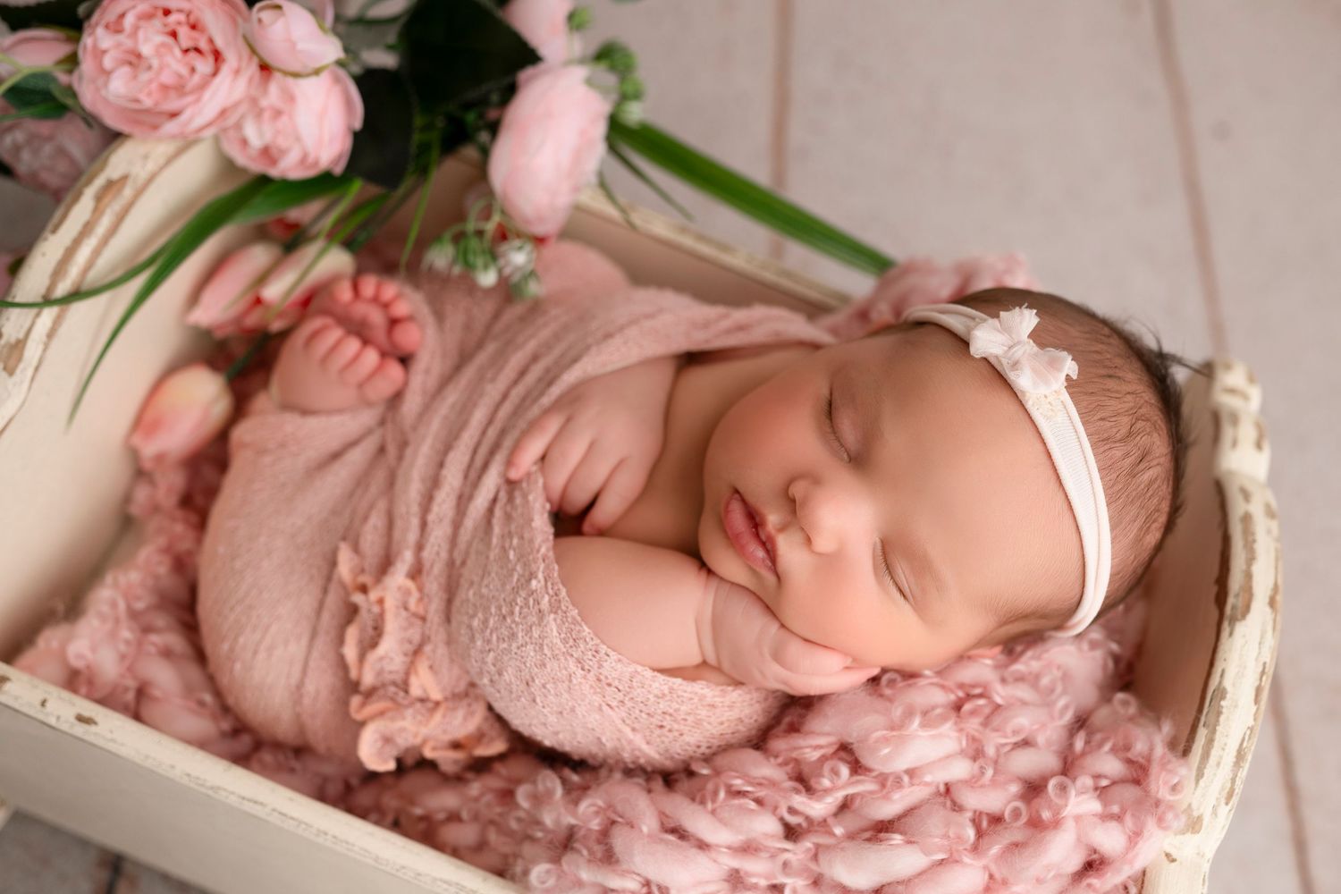 Sleeping newborn wrapped in soft pink fabric lying on textured blanket in white wooden crate with roses.