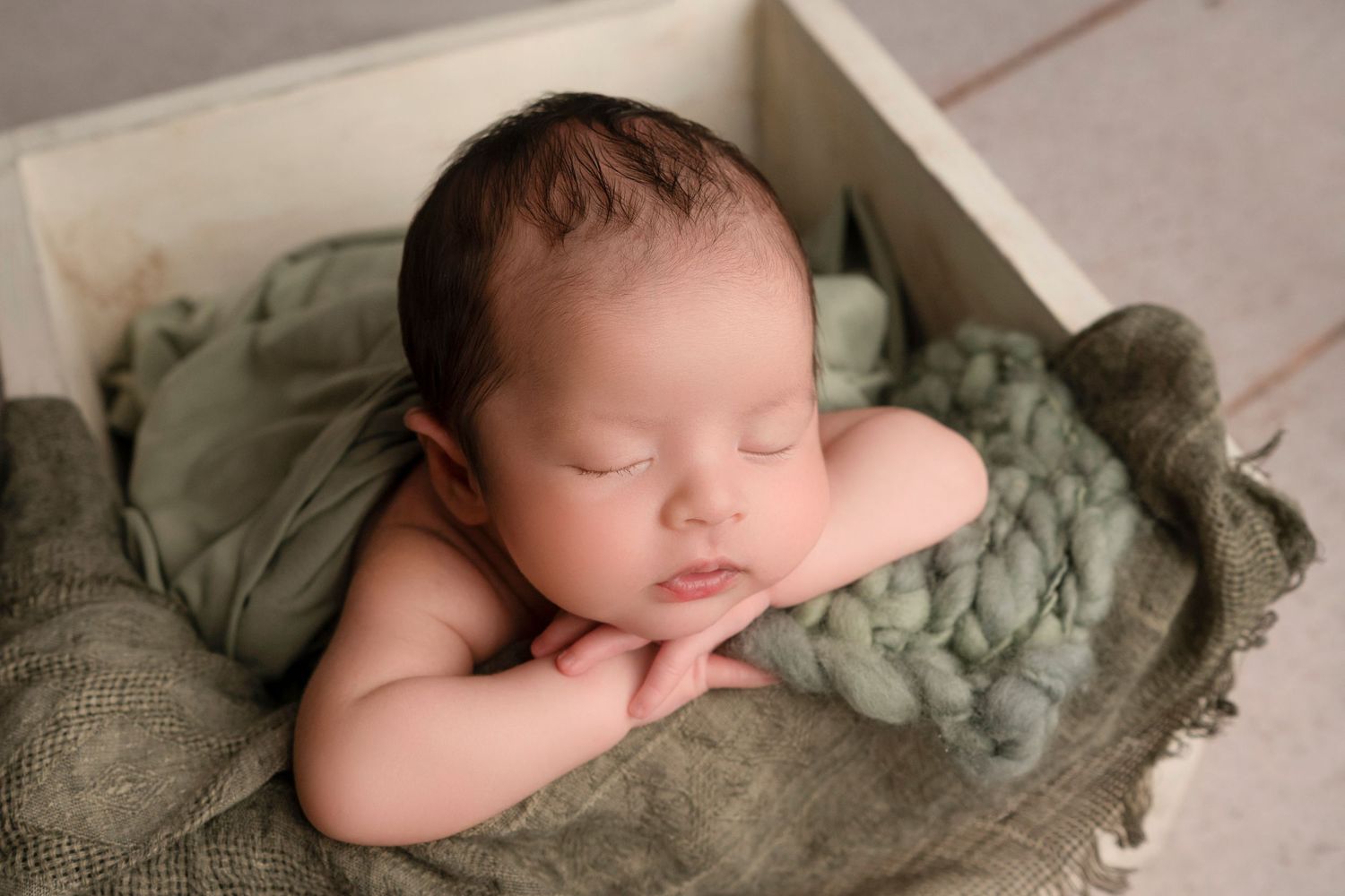 Sleeping newborn rests peacefully in wooden box with soft sage green blanket and wrap.