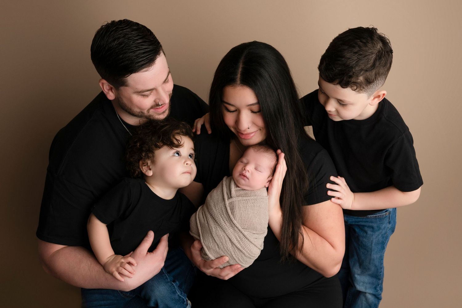 Family of five posing for newborn portrait, parents and two boys in black shirts cradling swaddled baby.