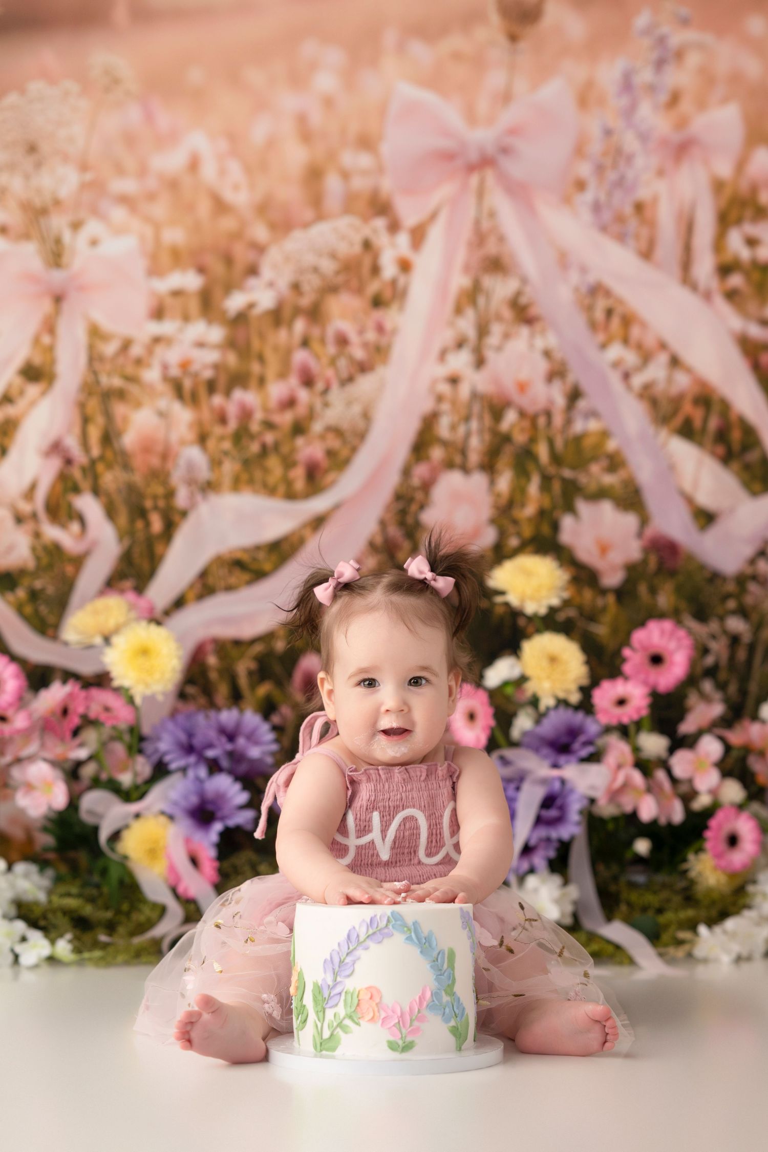 Baby girl in pink tutu sits before floral first birthday cake smash backdrop with colorful flowers and pink bows.
