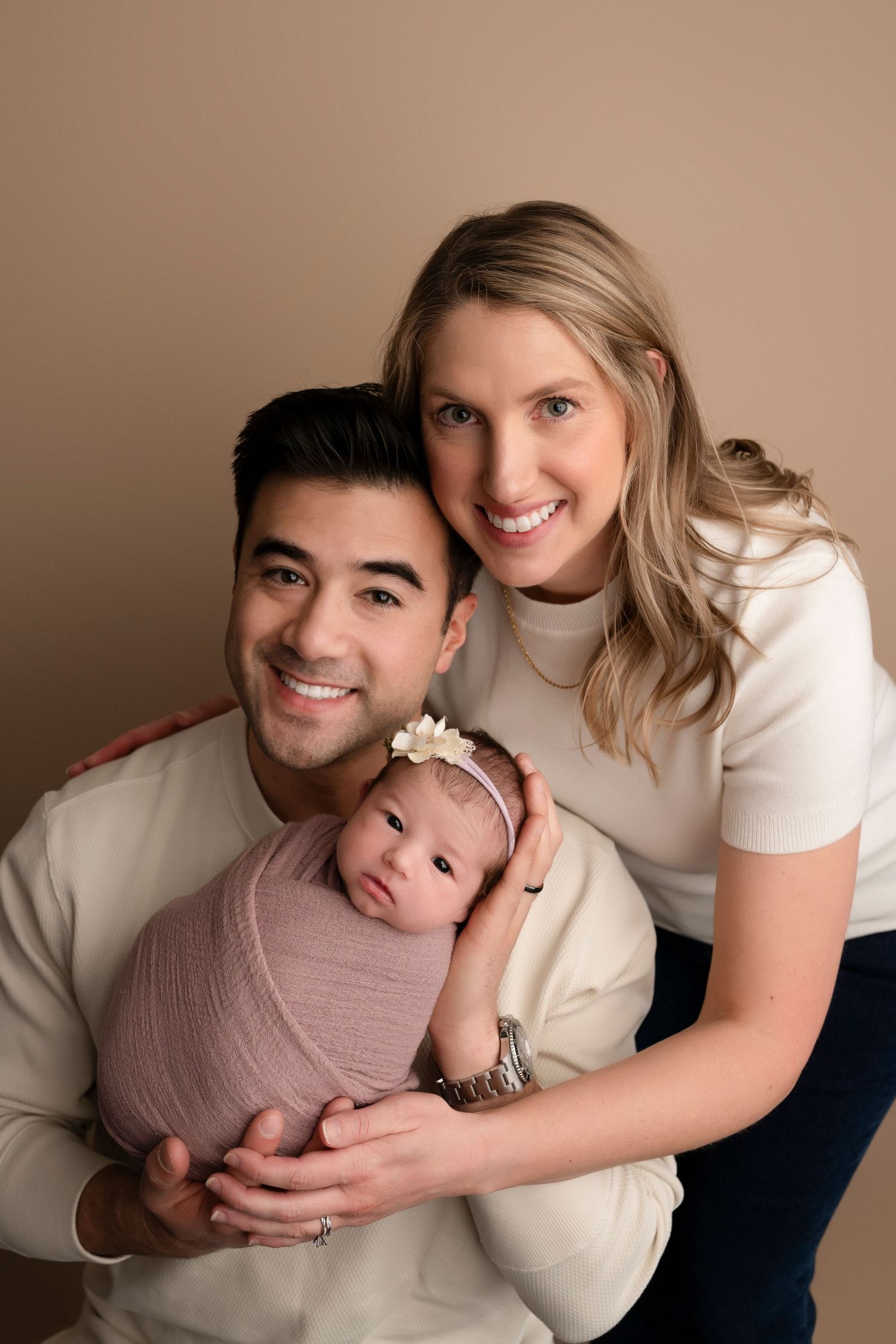 Happy couple in cream tops smiling with their newborn baby girl wrapped in pink, studio portrait.