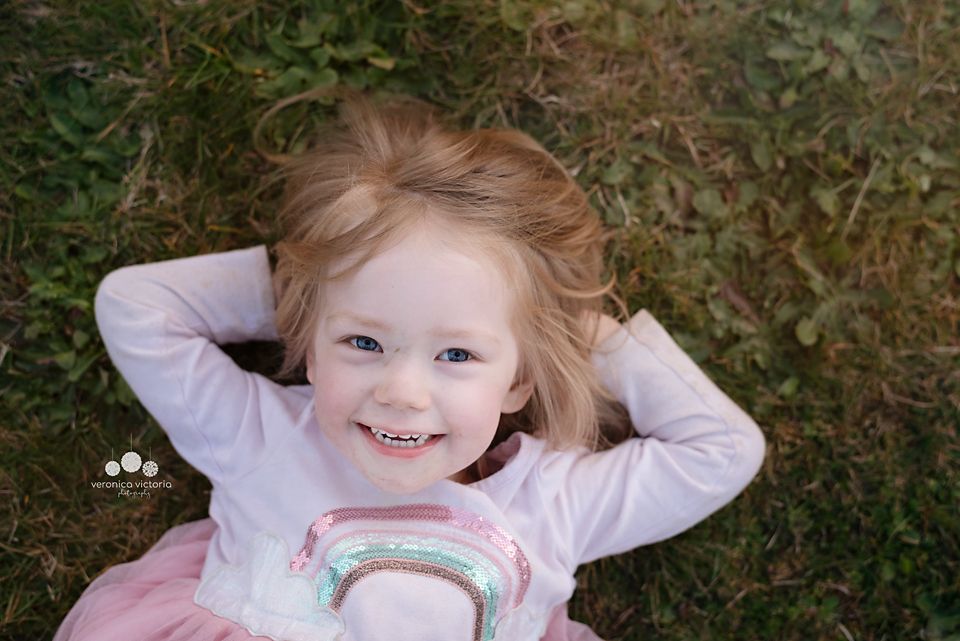 A smiling toddler girl with blonde hair lies on green grass, hands behind her head, wearing a pink rainbow shirt.