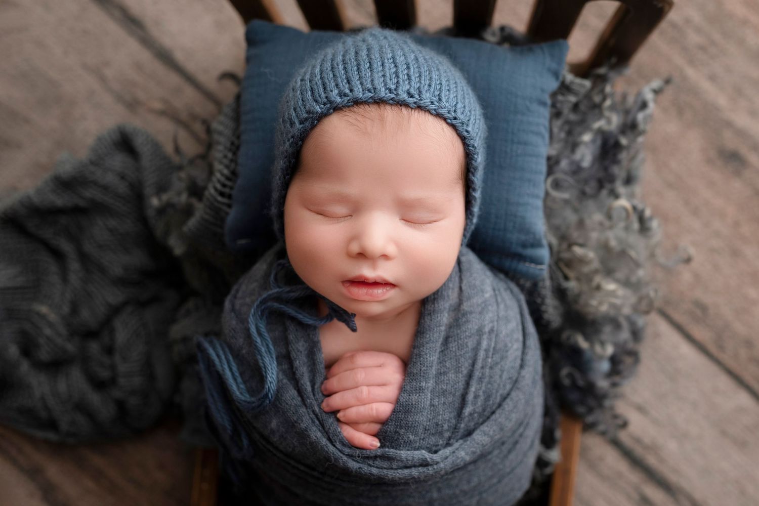 Sleeping newborn baby wrapped in gray knit blanket wearing blue bonnet, posed in wooden chair for professional photo.