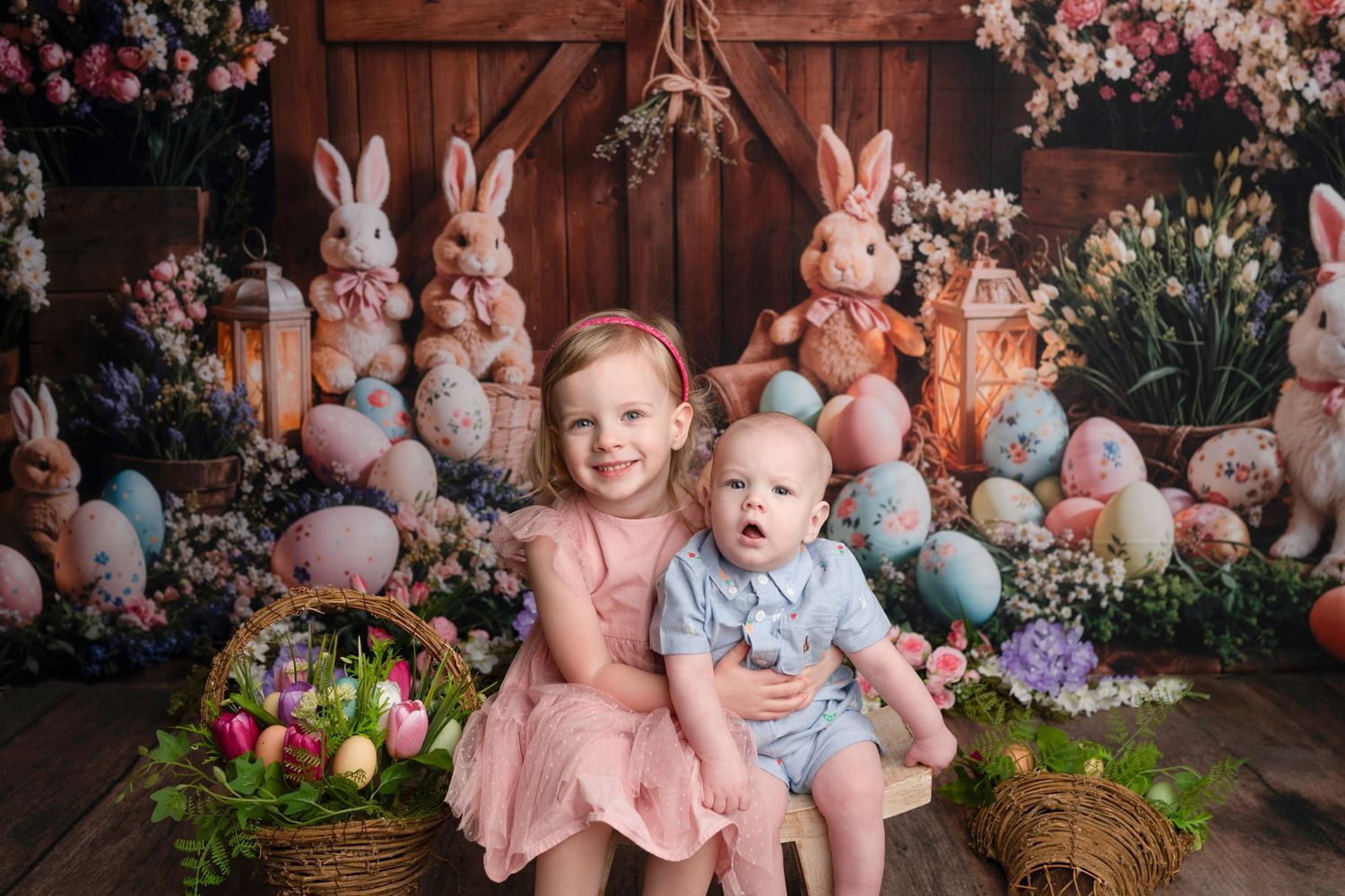 Two children pose with Easter eggs, bunnies, and spring flowers in a festive holiday photo backdrop setting.