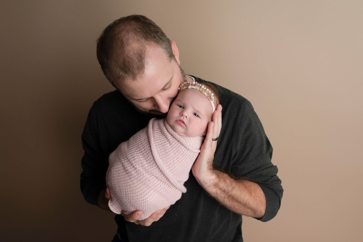 A bald father tenderly kisses his swaddled newborn baby girl in a pink wrap and headband against a neutral background.