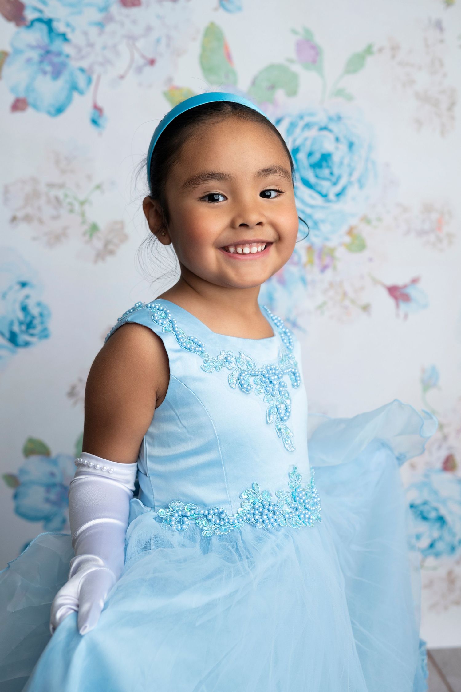 Young girl smiling in a light blue princess dress with white gloves and blue headband against floral backdrop.