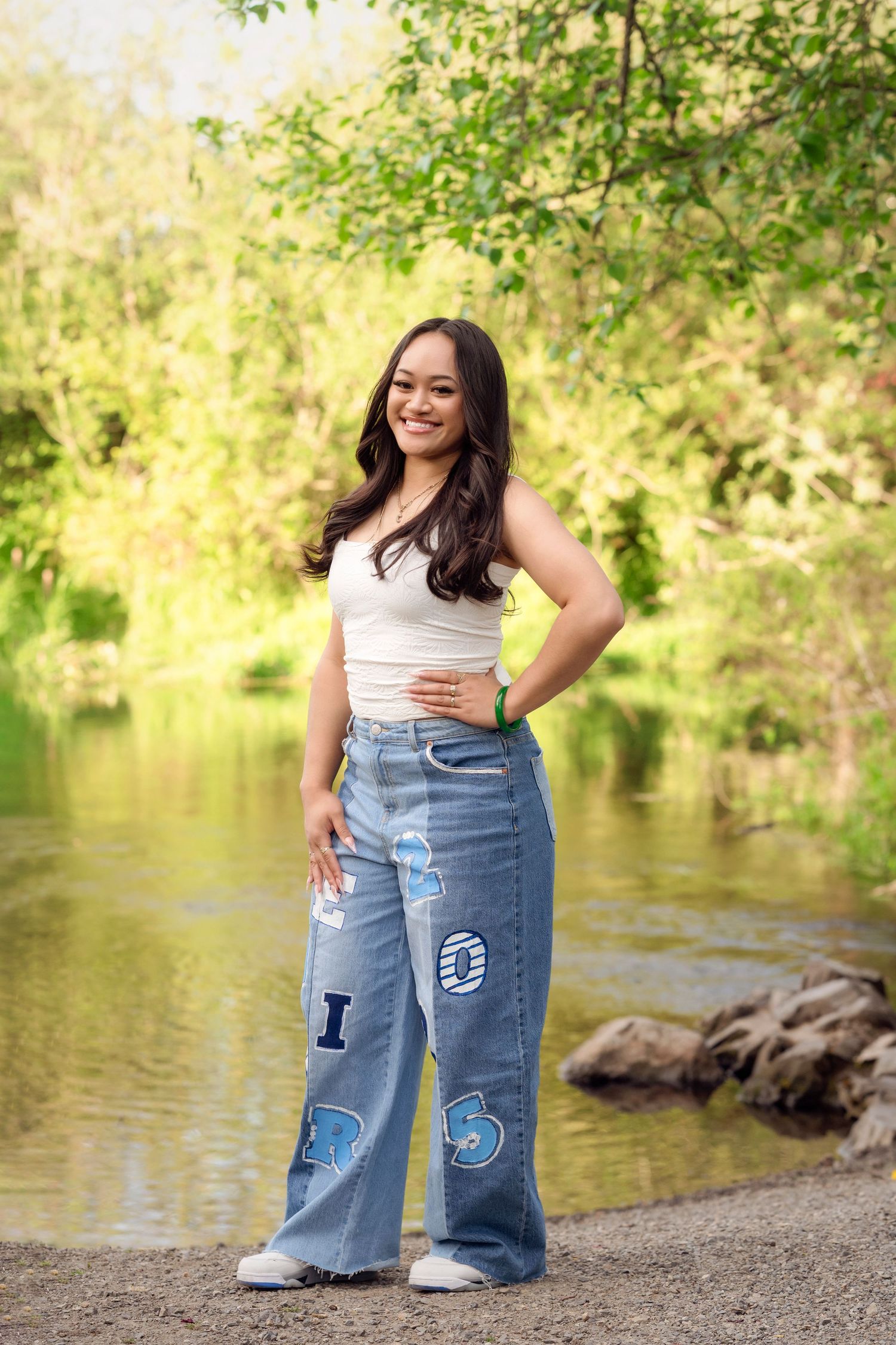 Person in white top and decorated jeans poses near a serene lake with trees in the background.
